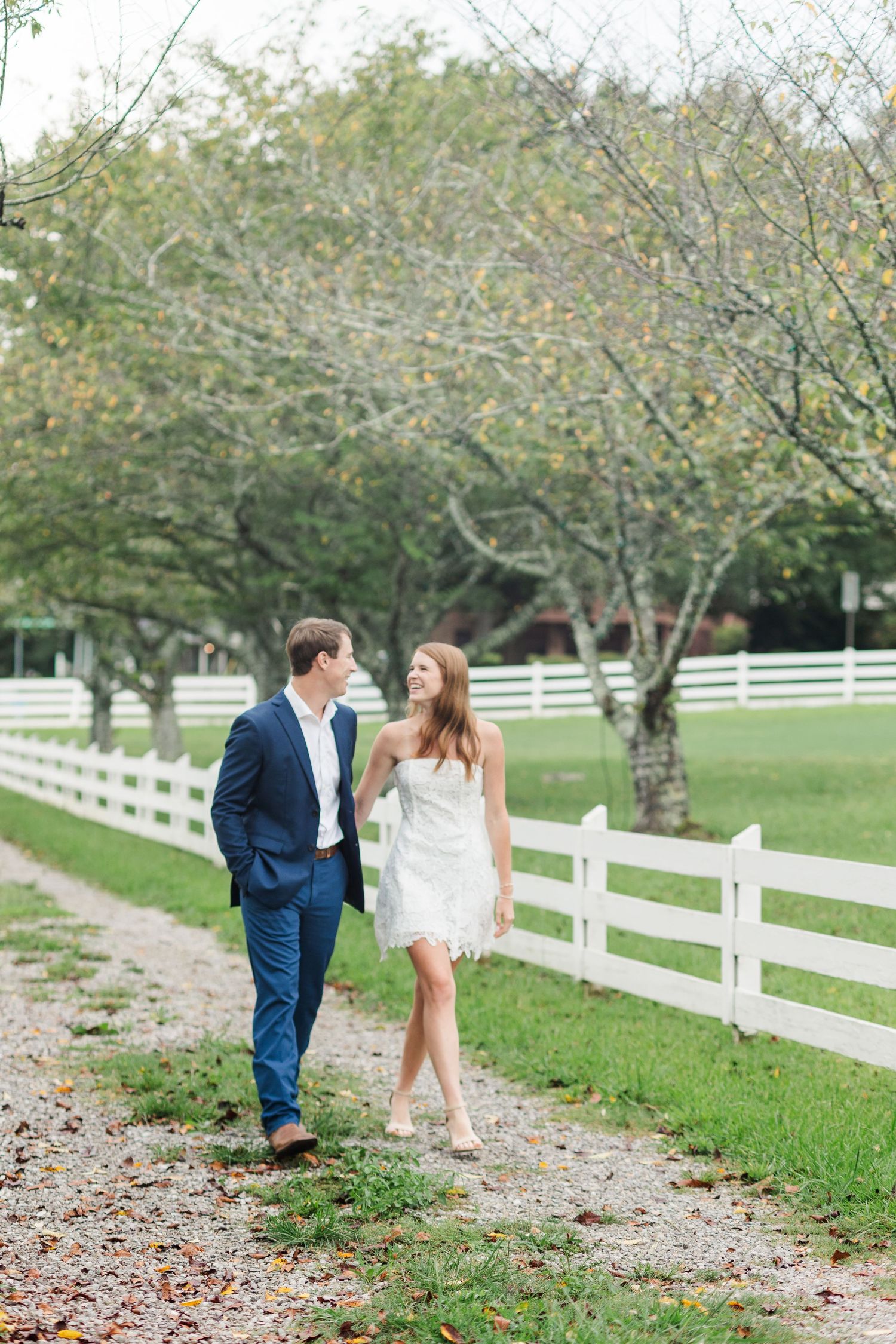 Couple walking along a gravel path by a white fence, man in navy suit, woman in white dress, lush green trees.