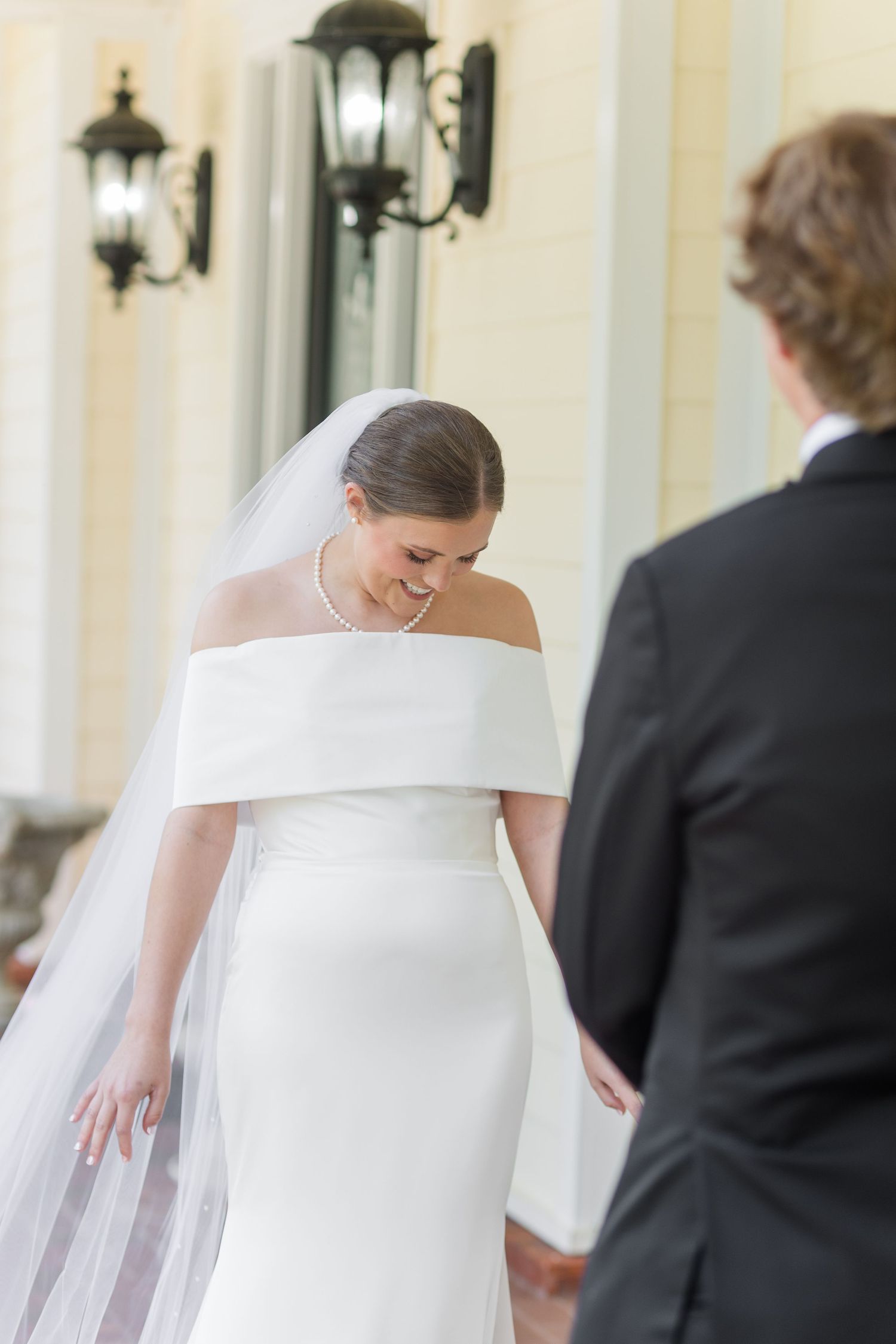 Blushing bride in white off-shoulder wedding dress laughs softly while holding grooms hand outdoors.