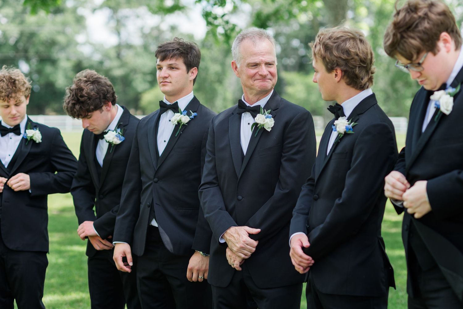 Groom and groomsmen in black tuxedos with white boutonnieres gathered outdoors before wedding.