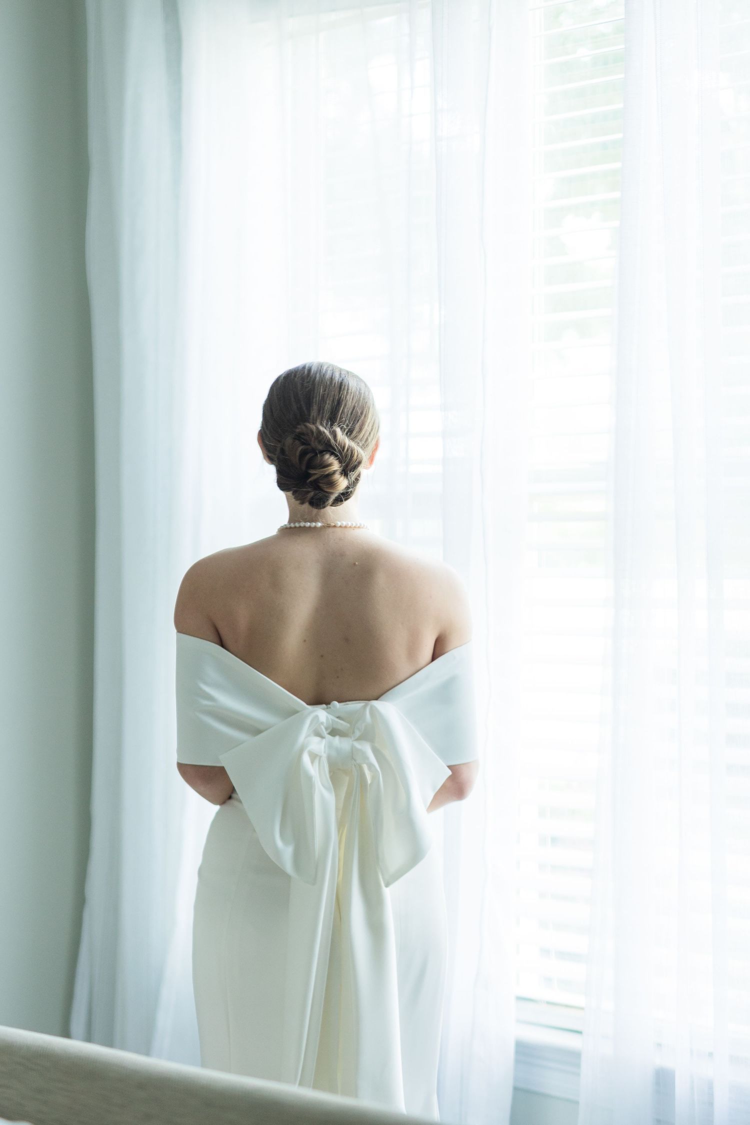 Bride in elegant off-shoulder white gown with large bow back stands before sheer curtained window.
