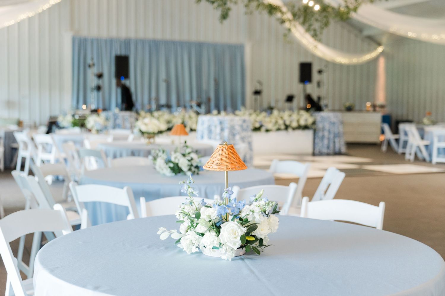 Soft blue and white wedding reception hall with round tables, floral centerpieces, and draped ceiling decorations.
