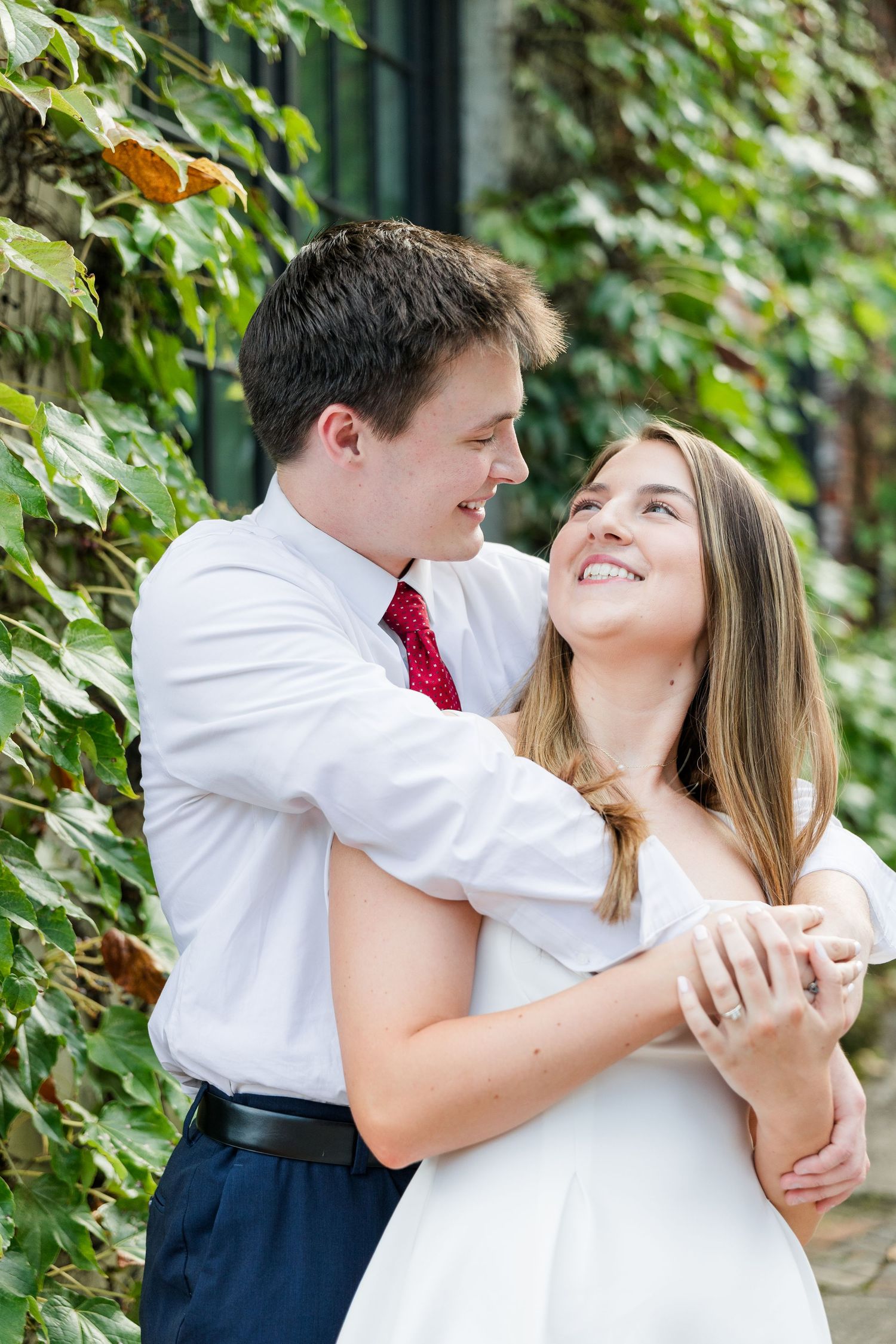 Happy couple embracing and smiling at each other in front of lush green ivy-covered wall outdoors.