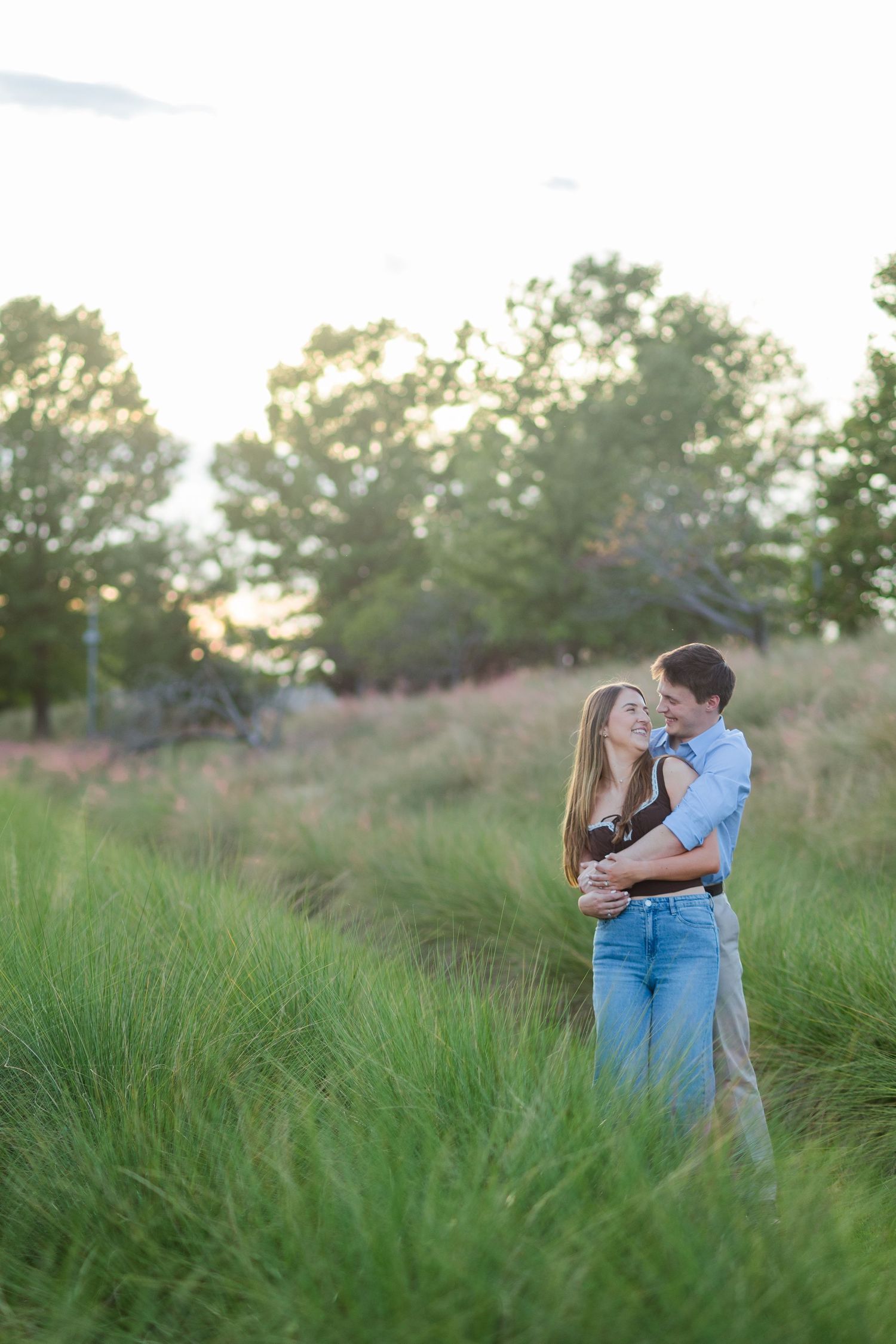 Romantic couple embracing in tall green grass field at golden hour with trees in background.