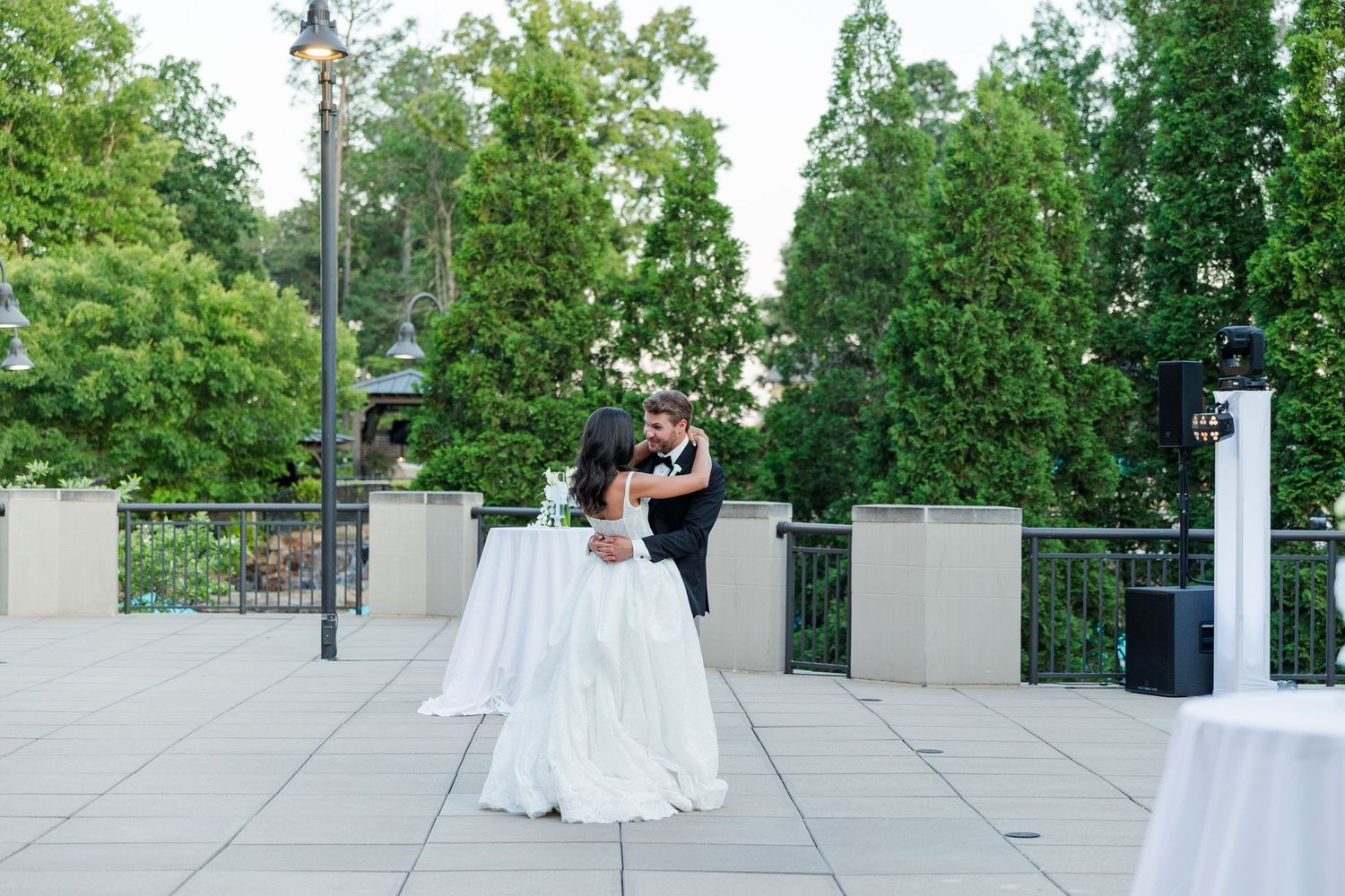 A bride and groom share a romantic dance on an outdoor terrace surrounded by lush green trees at their wedding reception.