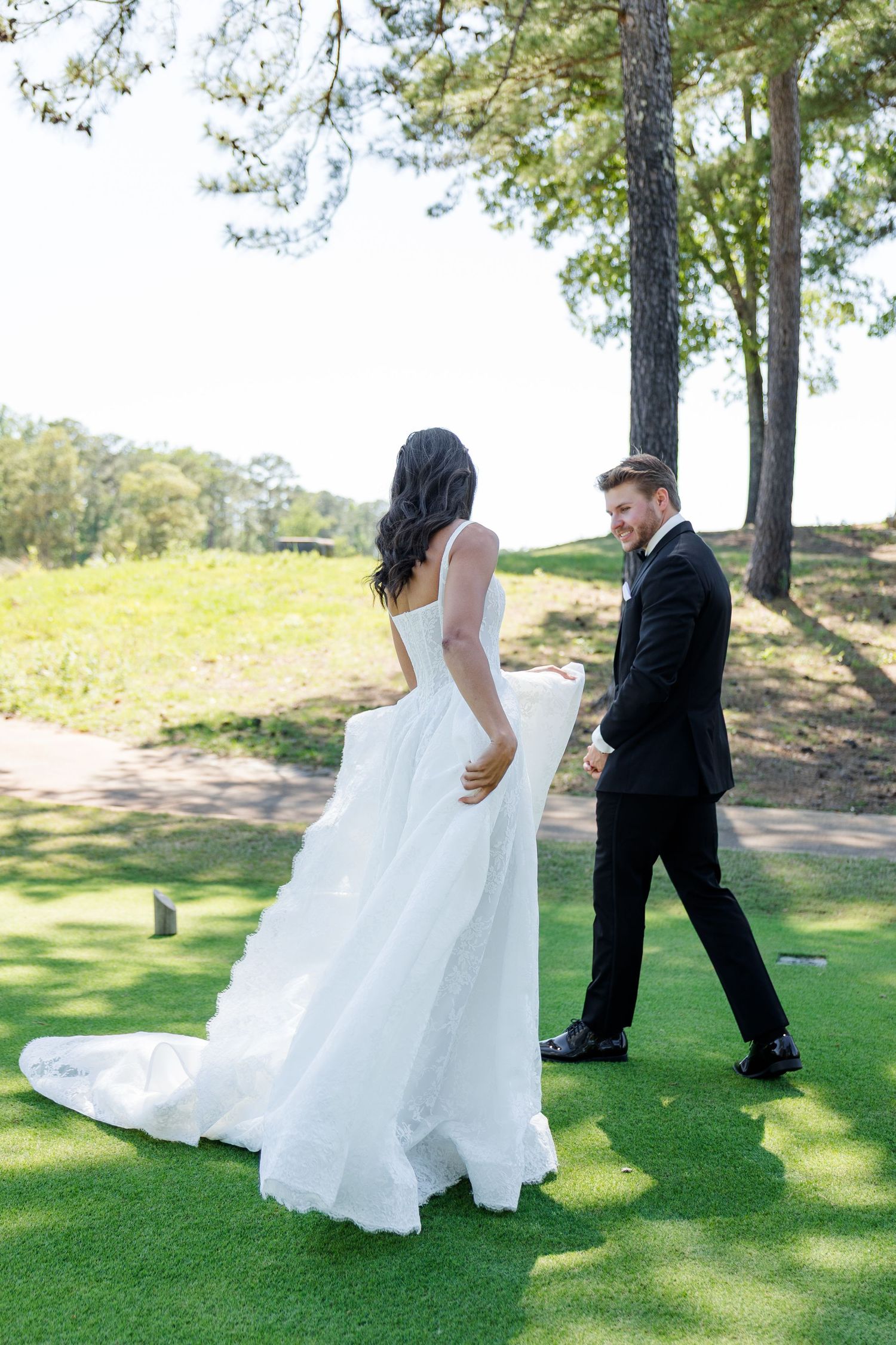 A bride in a white gown and groom in a black suit walk hand in hand on a lush green golf course.