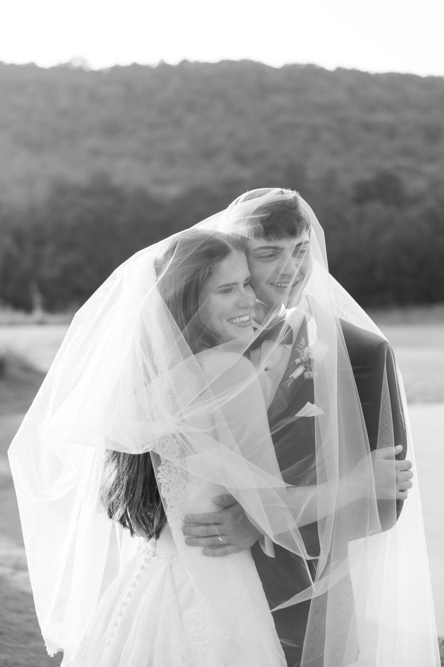 Black and white photo of bride and groom sharing a tender moment under a flowing veil outdoors.