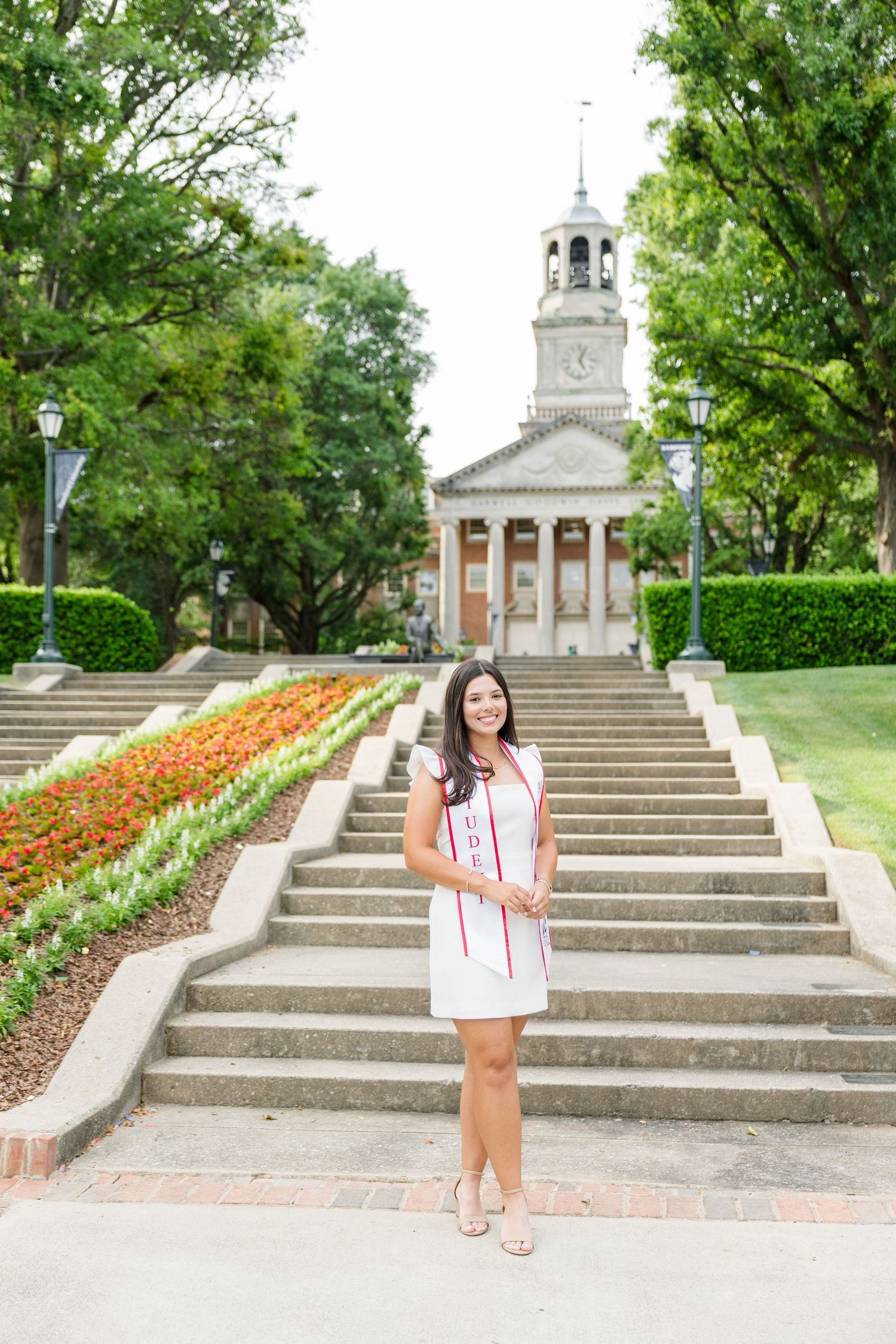 Female graduate in white dress with student athlete sash poses on university steps with historic bell tower building.