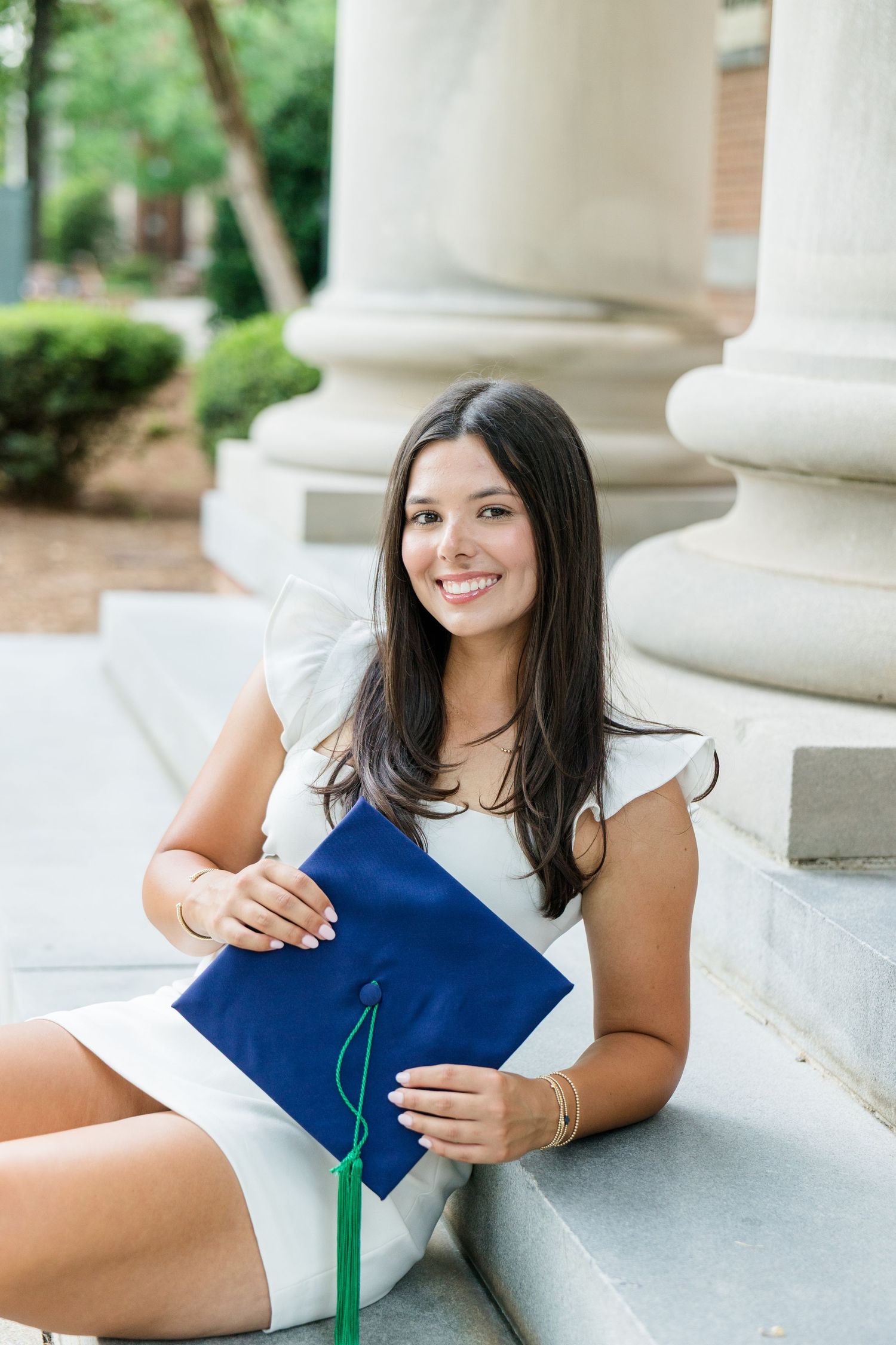 Smiling female graduate sitting on steps holding a blue graduation cap with green tassel outdoors.
