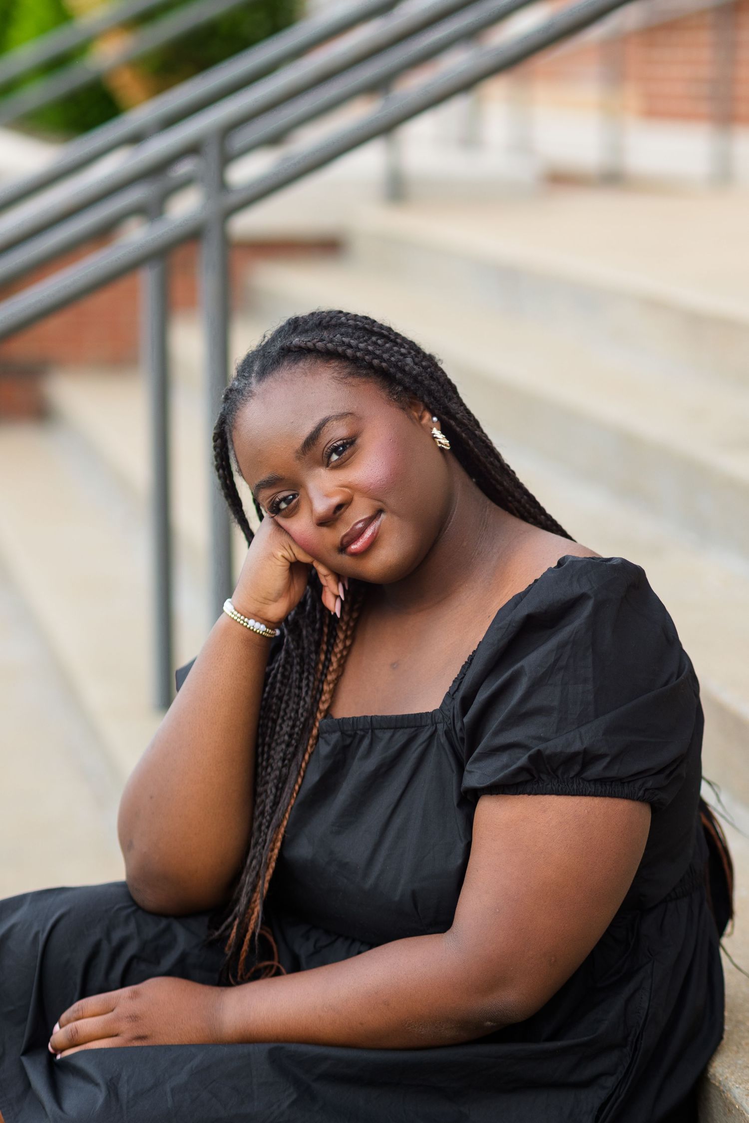 Young Black woman with long braids in black dress sitting on steps, resting head on hand.