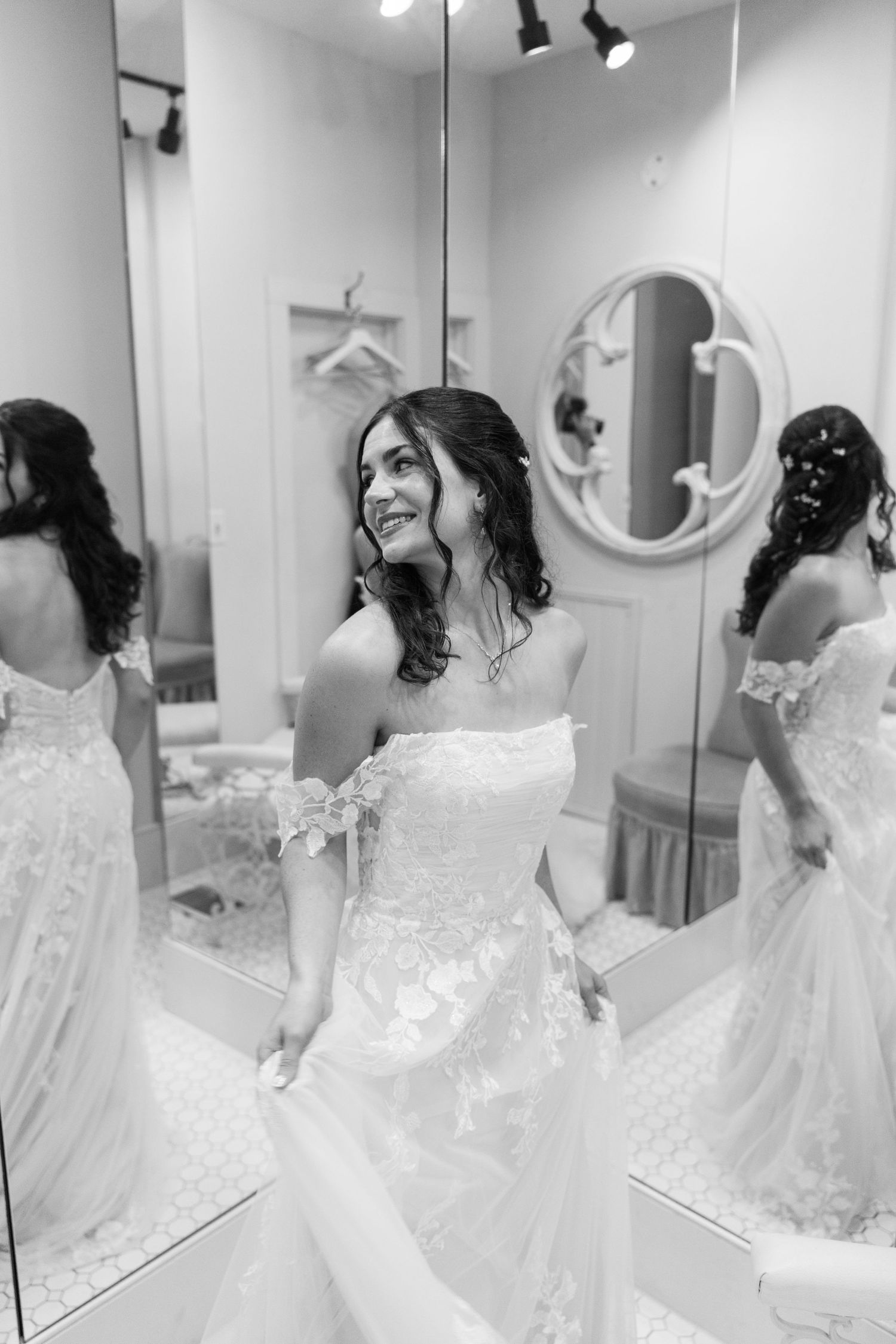 Black and white photo of a bride in an off-shoulder lace wedding gown smiling in a bridal boutique mirror.