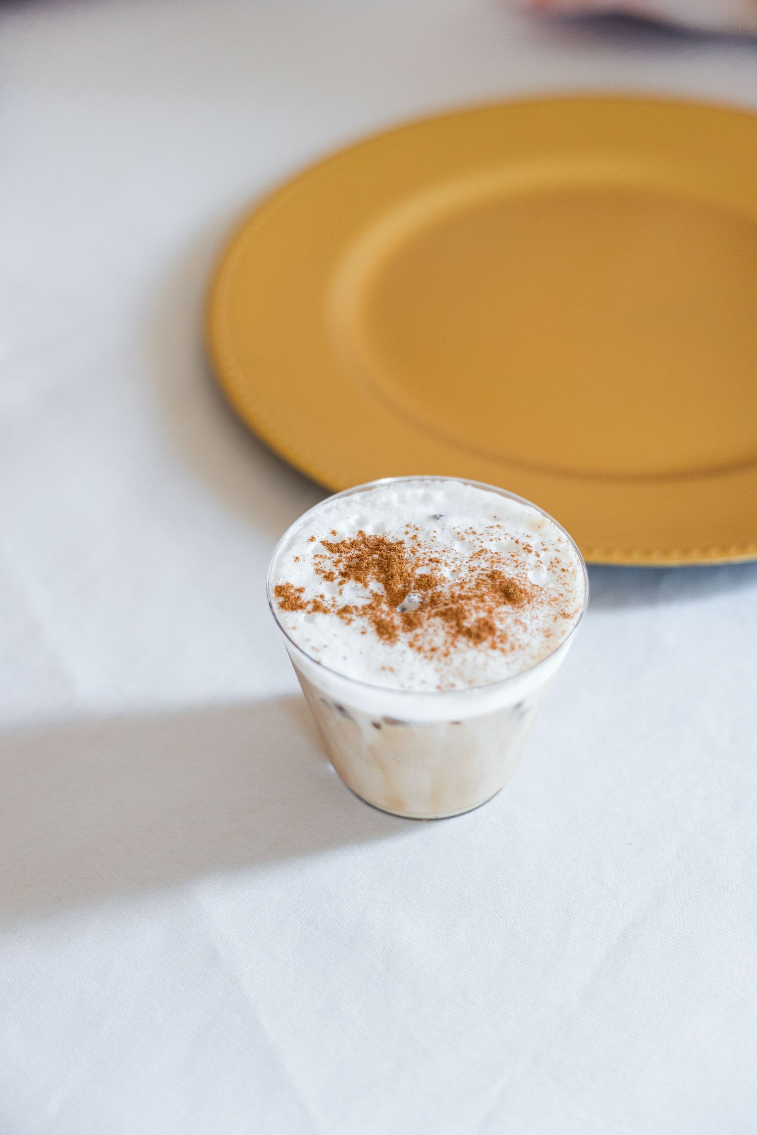 A frothy iced latte topped with cinnamon powder in a clear glass beside a gold plate on a white tablecloth.