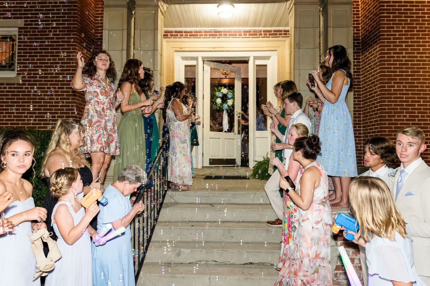 Wedding guests line steps blowing bubbles to celebrate the newlyweds outside a brick church entrance at night.