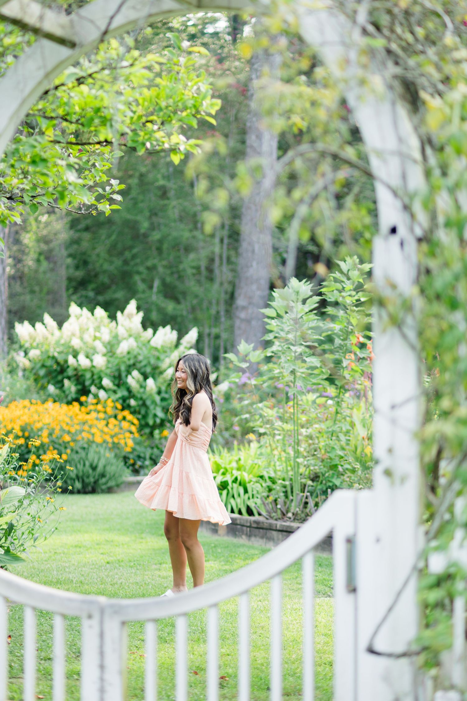 Young woman in pink dress standing in lush garden viewed through white arbor gate.