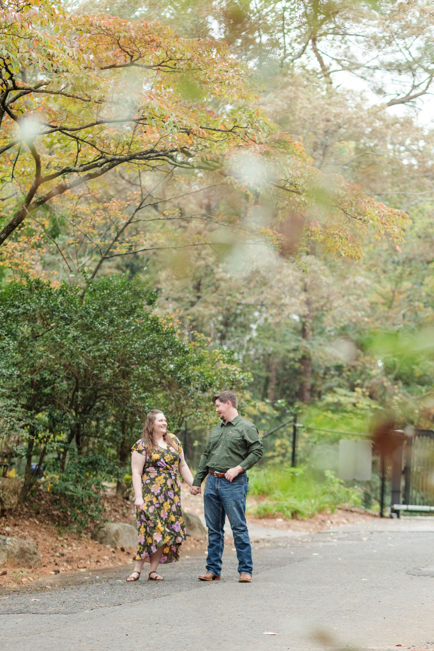 Couple holding hands in a park during fall, woman in floral dress, man in green shirt and jeans, surrounded by autumn foliage.