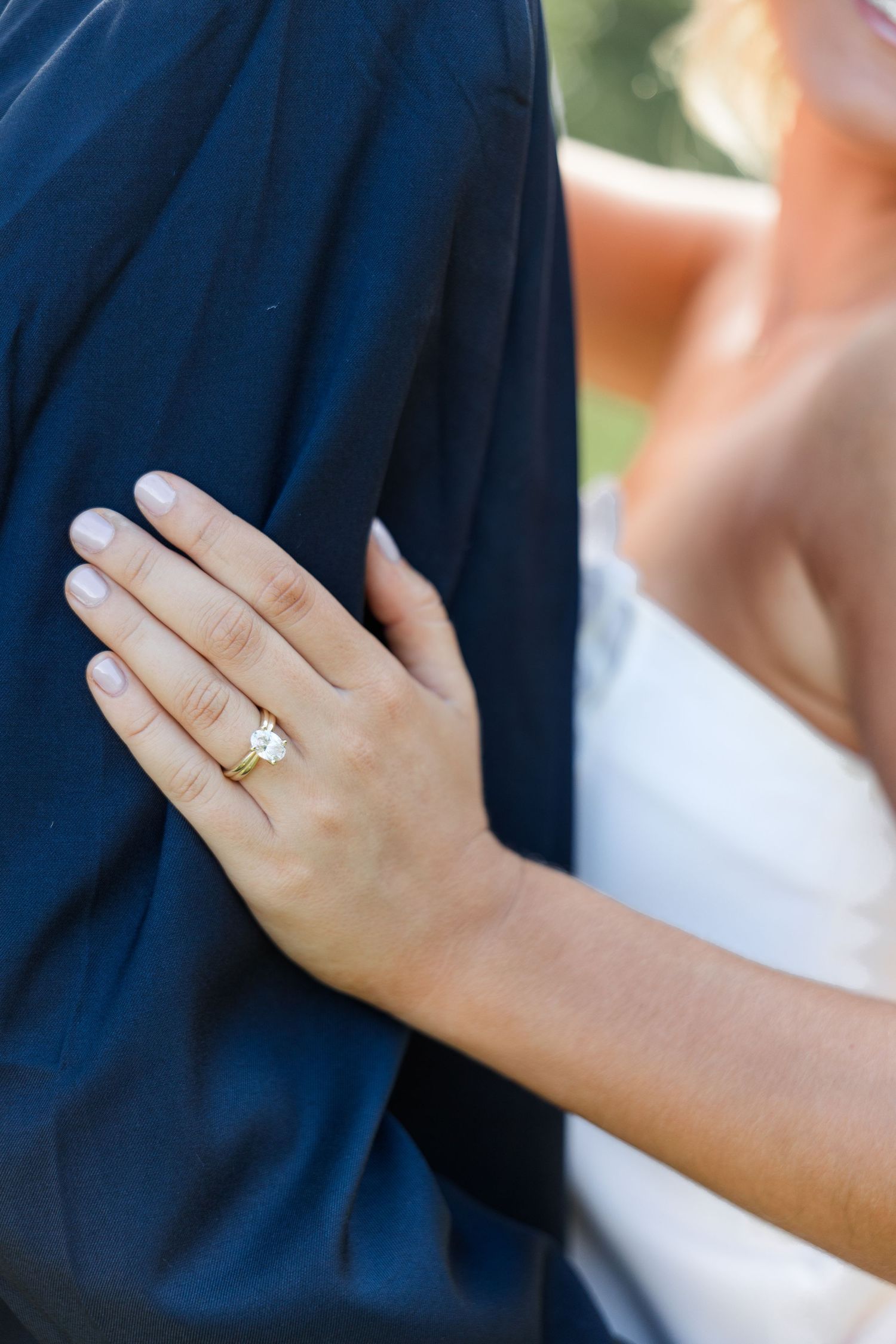 Close-up of a bride's hand with an oval diamond engagement ring on a gold band resting on groom's navy suit.