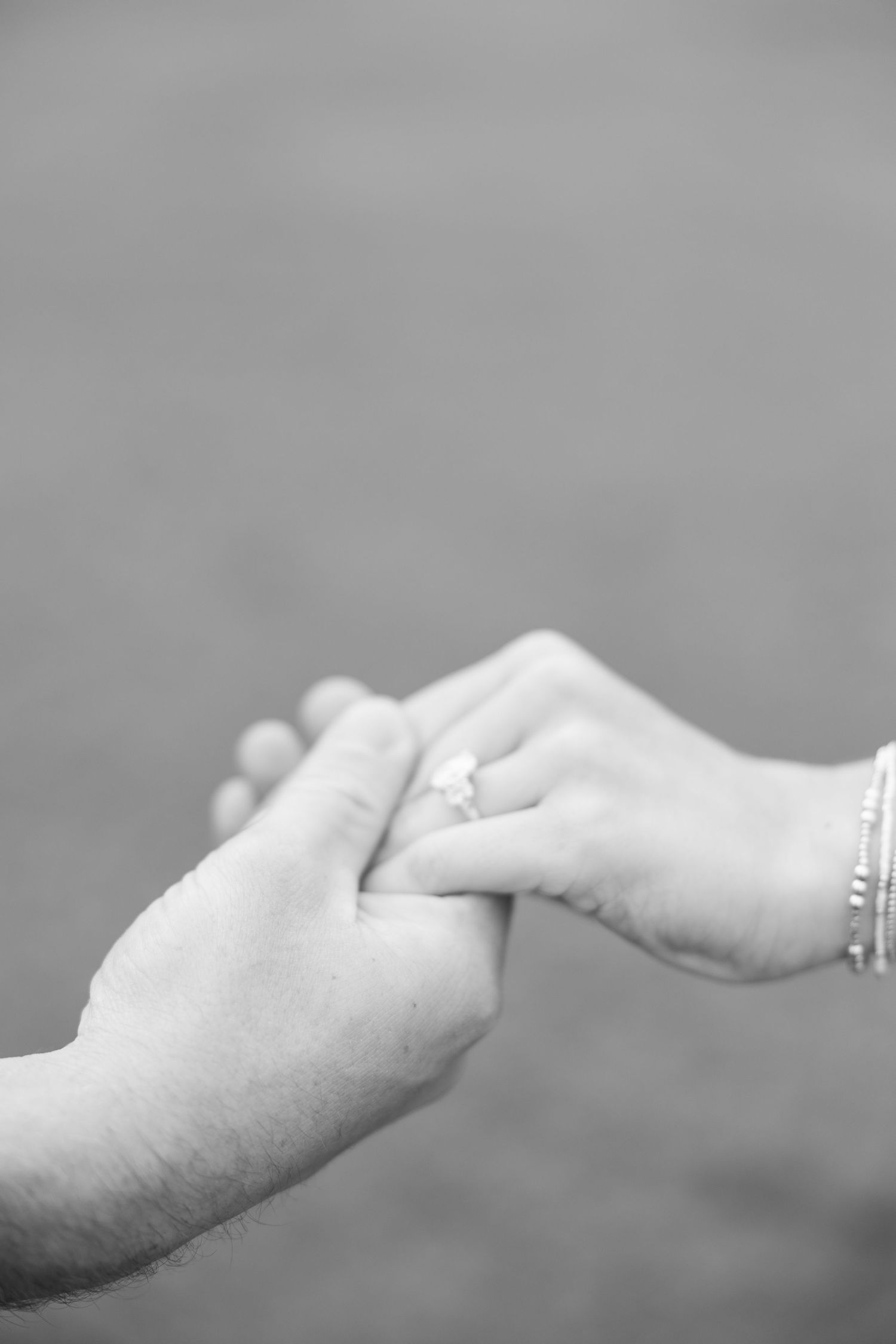 Black and white close-up of two hands clasped together, showing a sparkling engagement ring and a beaded bracelet.