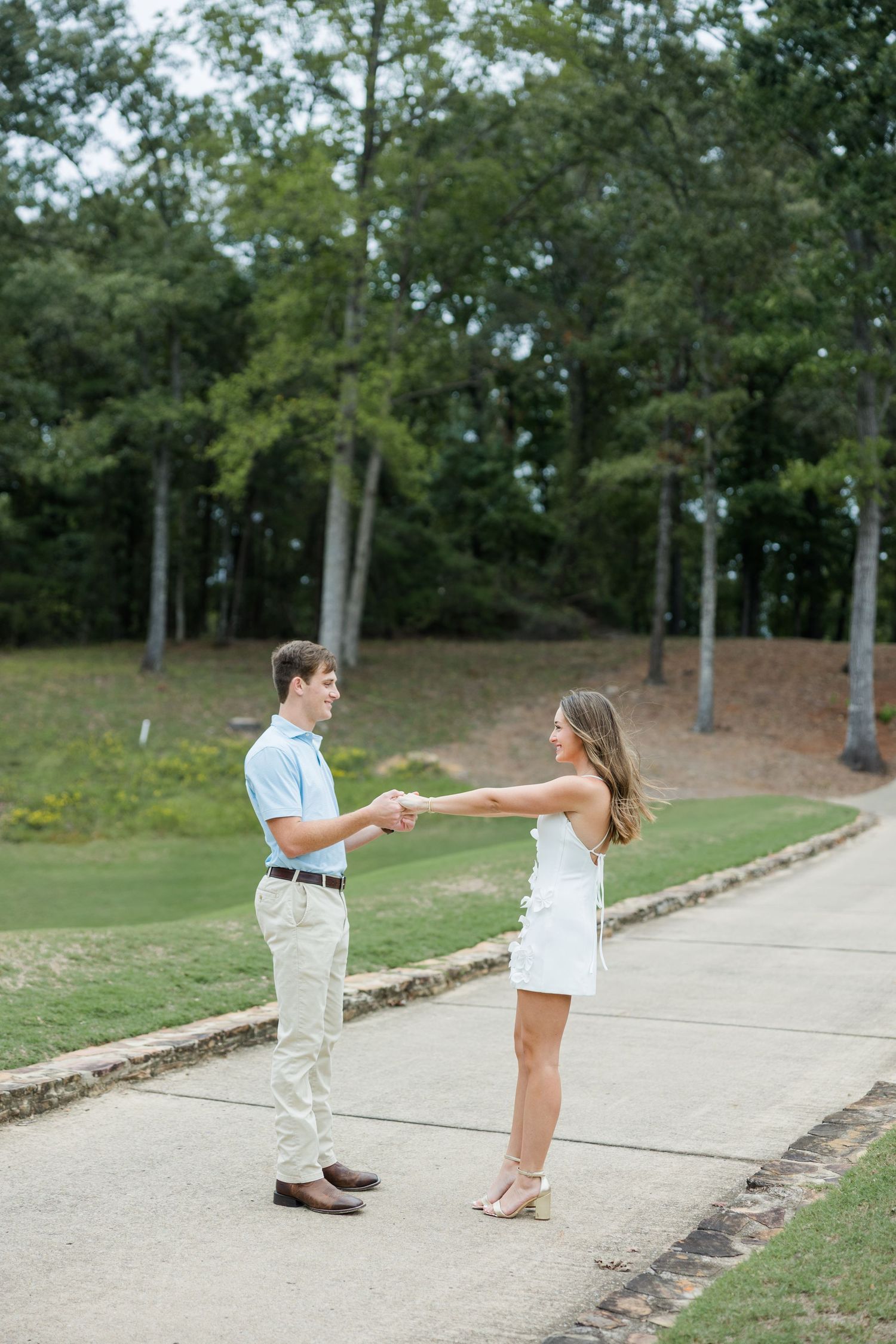 Smiling couple holding hands and facing each other on a paved path surrounded by green trees and grass.