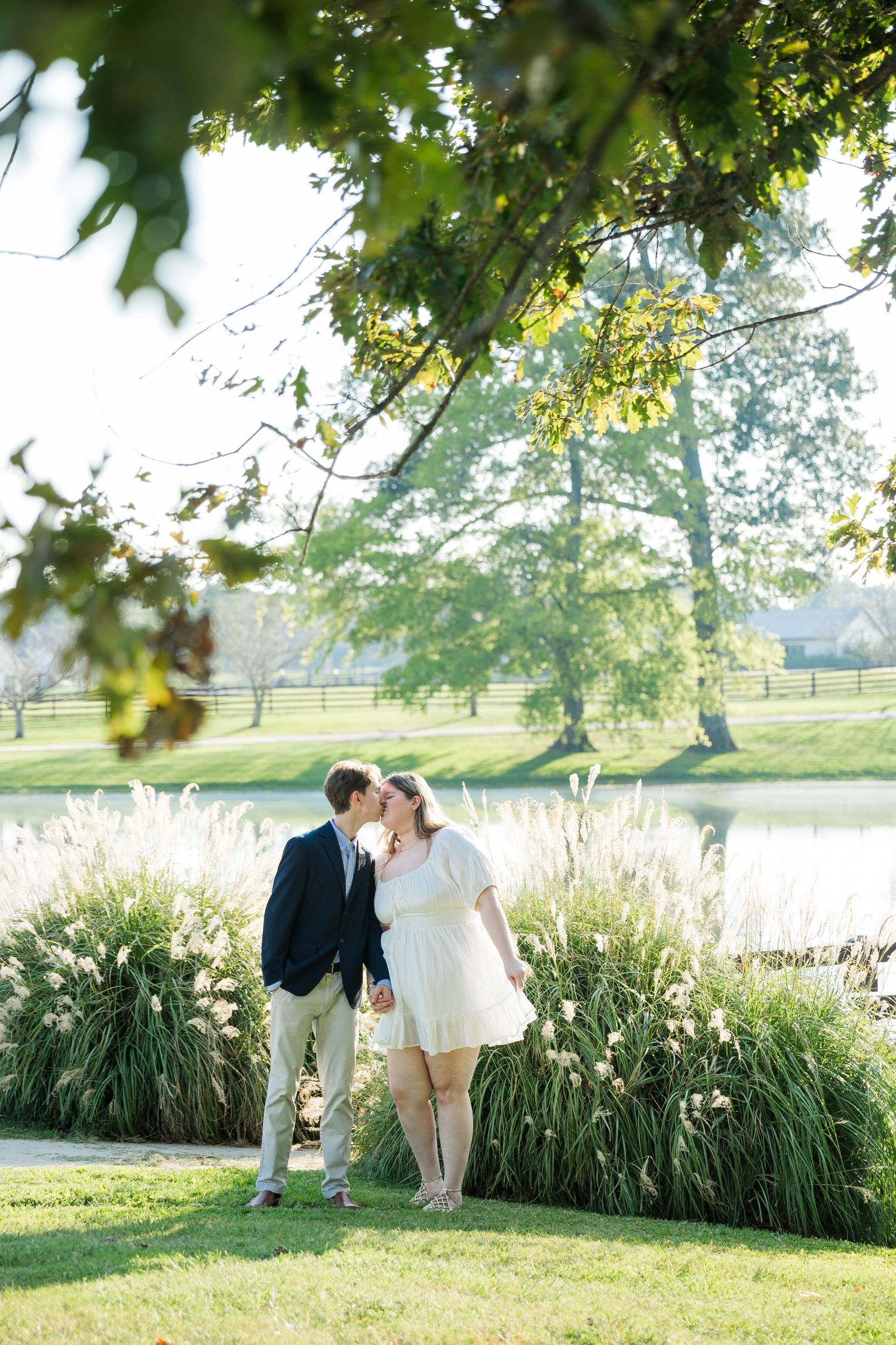 Couple sharing a kiss outdoors near a pond, surrounded by tall grasses and trees in a lush green park setting.