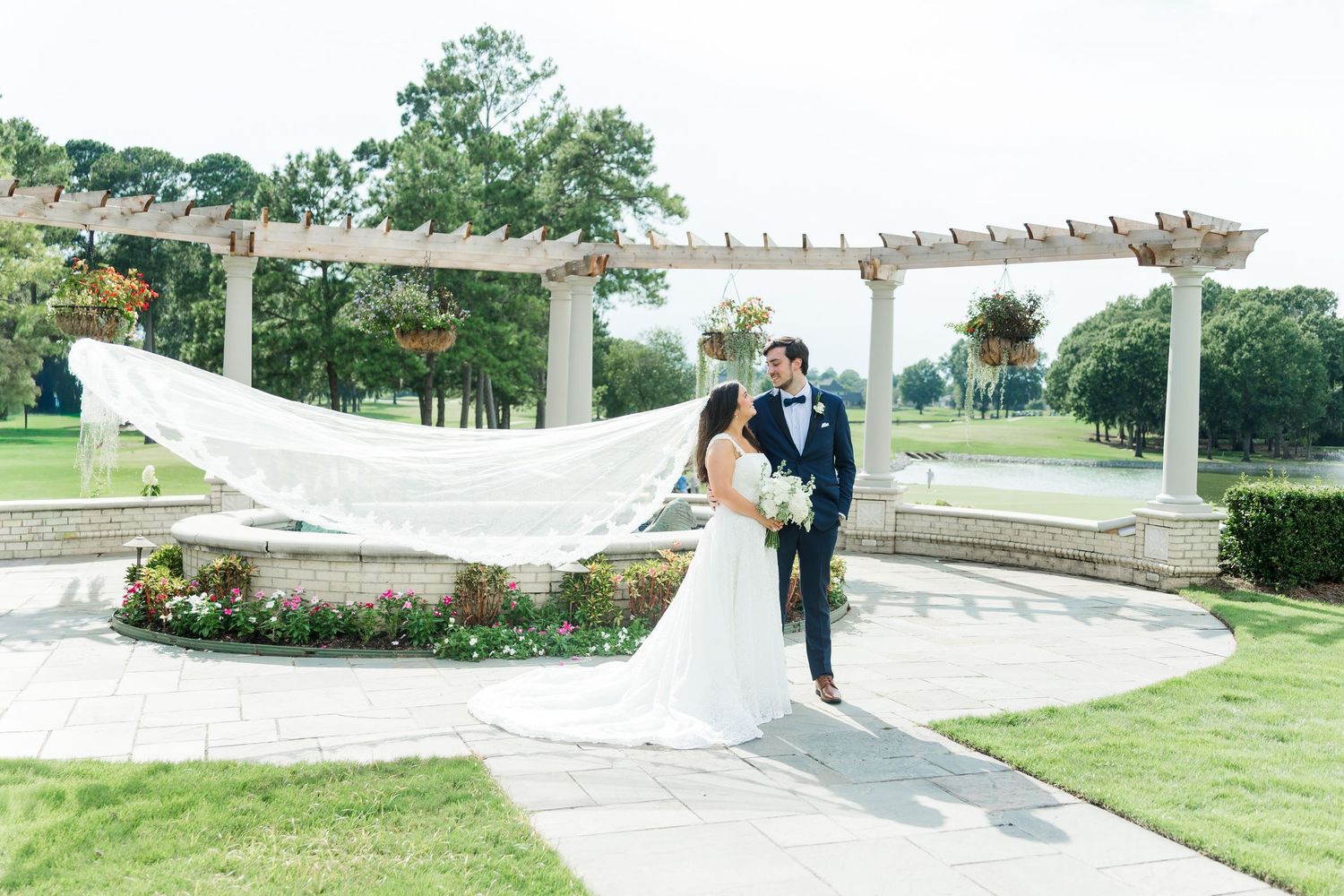 Bride and groom pose in garden as flowing veil billows in the wind at outdoor wedding.