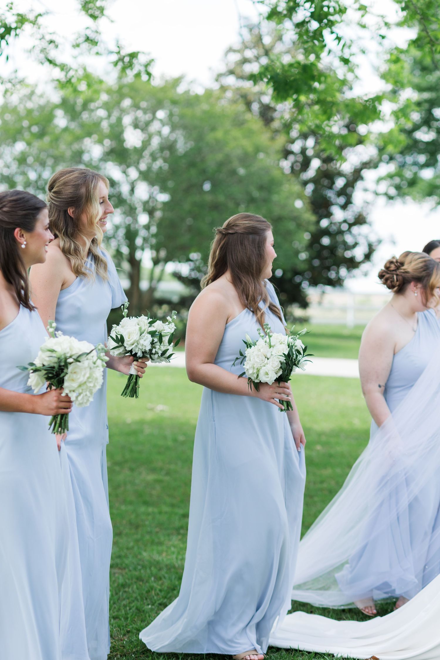 Bridesmaids in dusty blue chiffon gowns holding white floral bouquets stand outdoors on wedding day.
