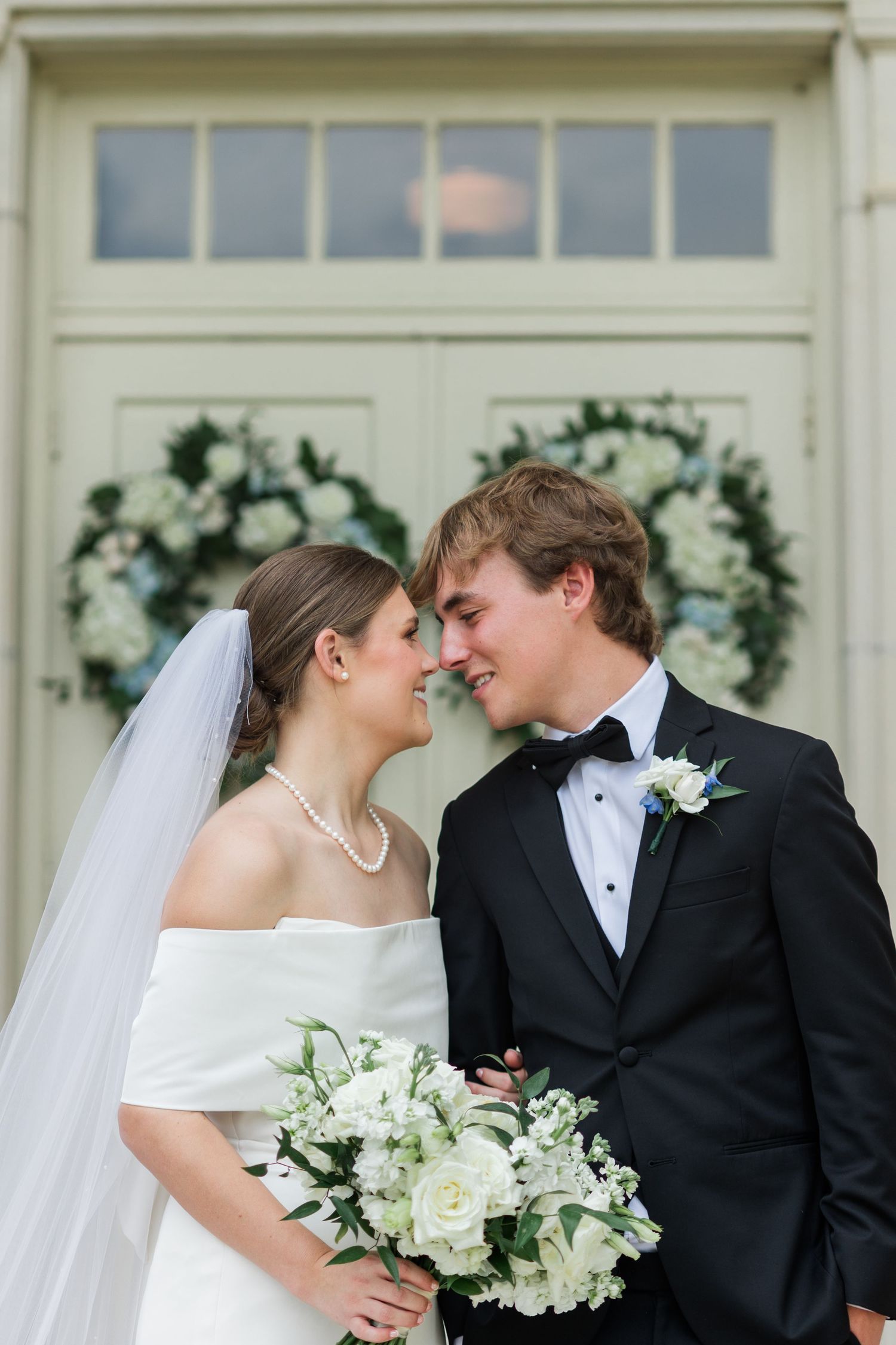 Bride and groom sharing an intimate moment before white doors with floral wreaths, bride wearing pearl necklace and veil.