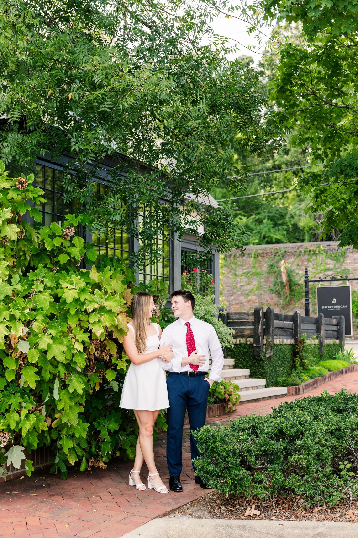 An engaged couple holds hands and smiles in a lush green garden, man in white shirt and red tie, woman in white dress.