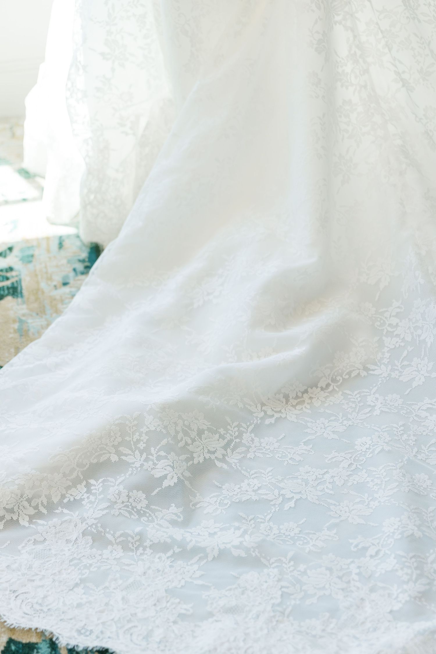 Close-up of an elegant white lace wedding dress with delicate floral embroidery draped gracefully near a bright window.