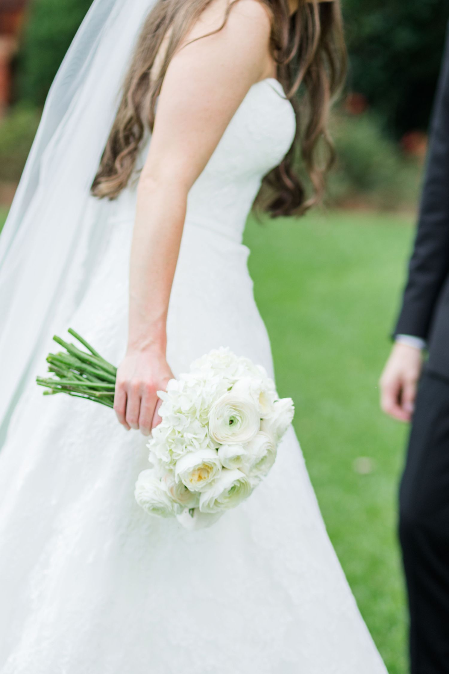 Bride in white strapless gown holding elegant white ranunculus and hydrangea wedding bouquet beside groom in black suit.