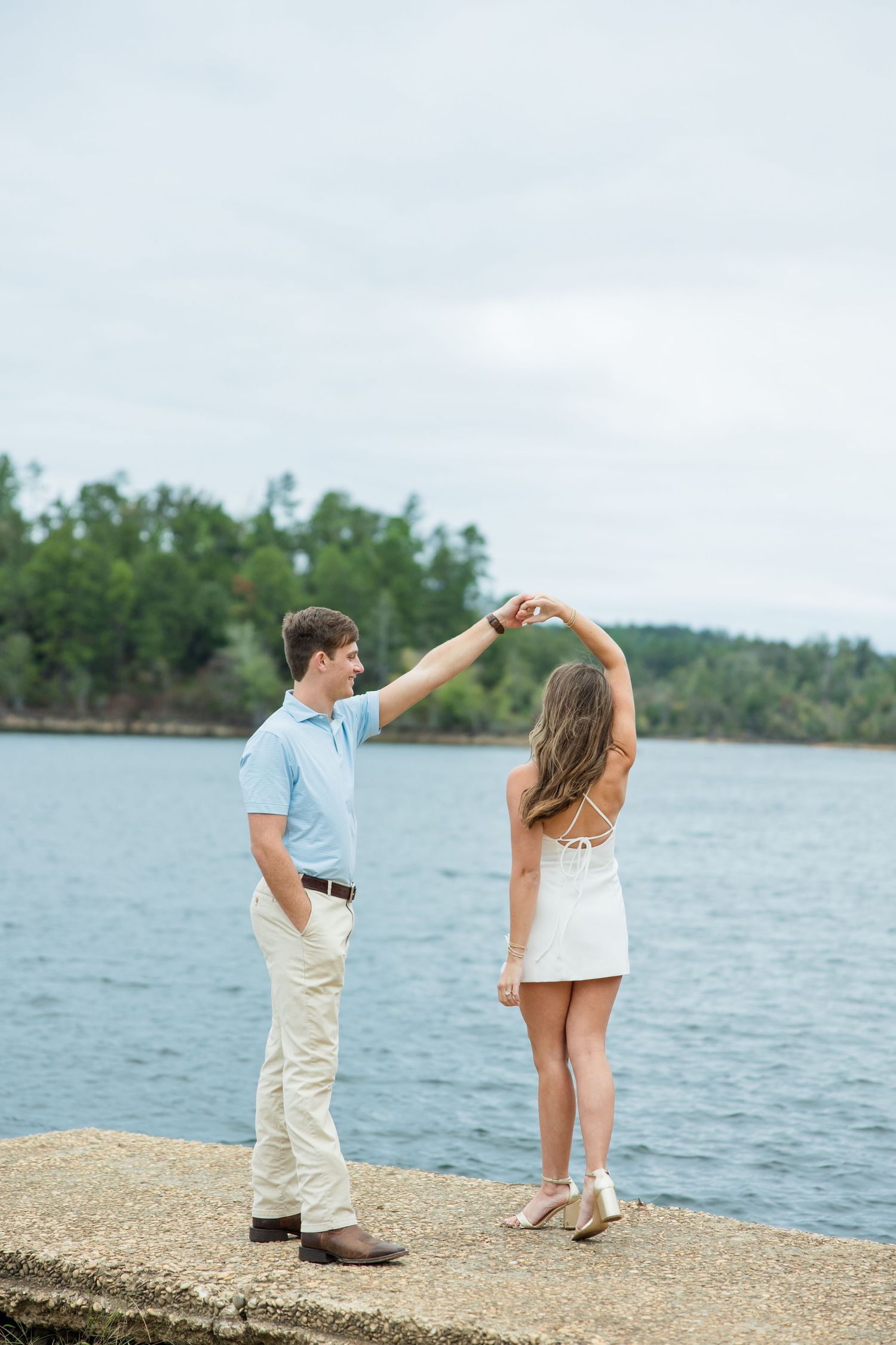 Couple in casual attire dancing on a stone pier as he twirls her beside a calm lake with a tree-lined shore.