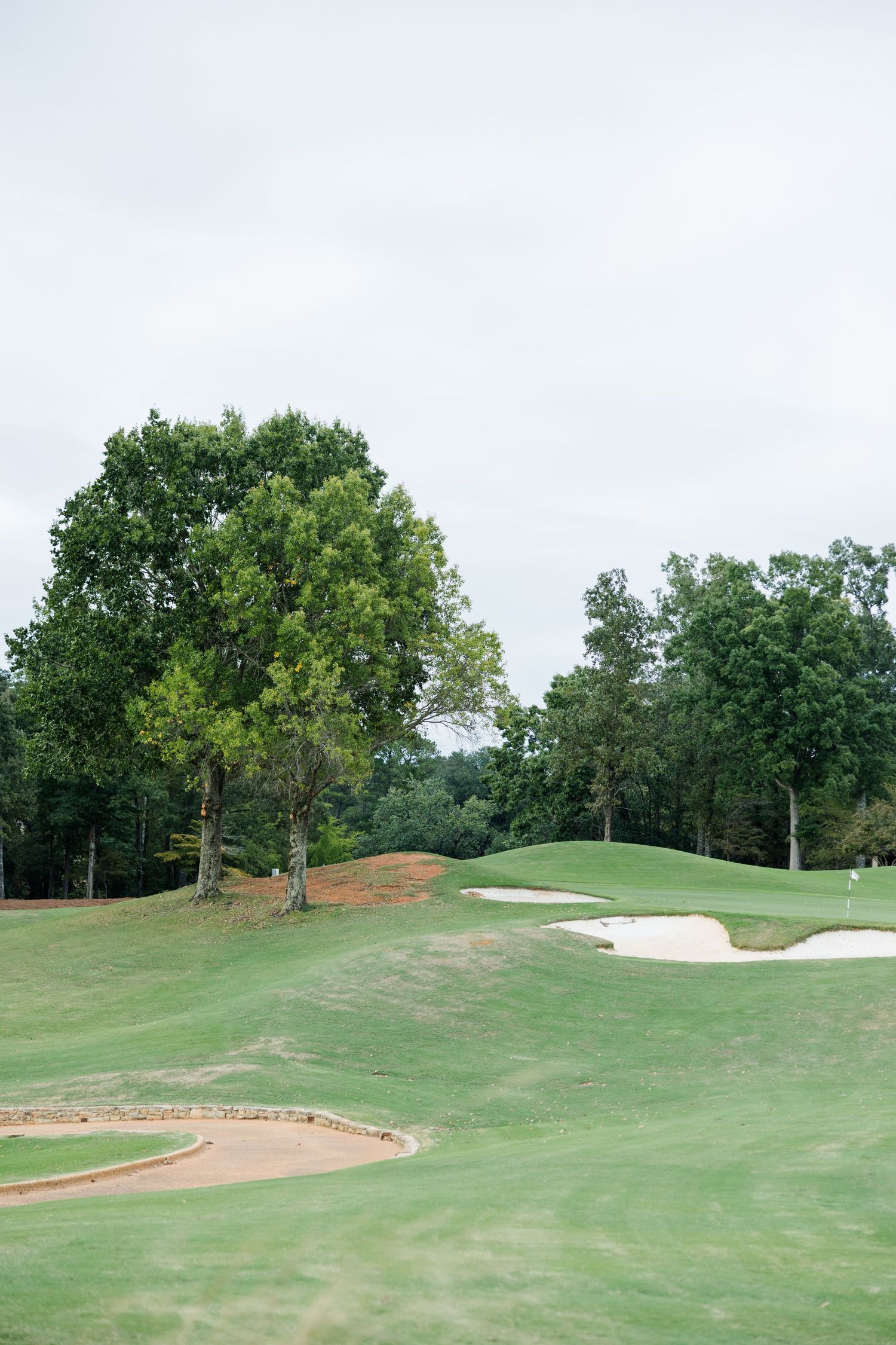 Golf course fairway with sand bunkers, rolling green hills and tall leafy trees under a cloudy sky.