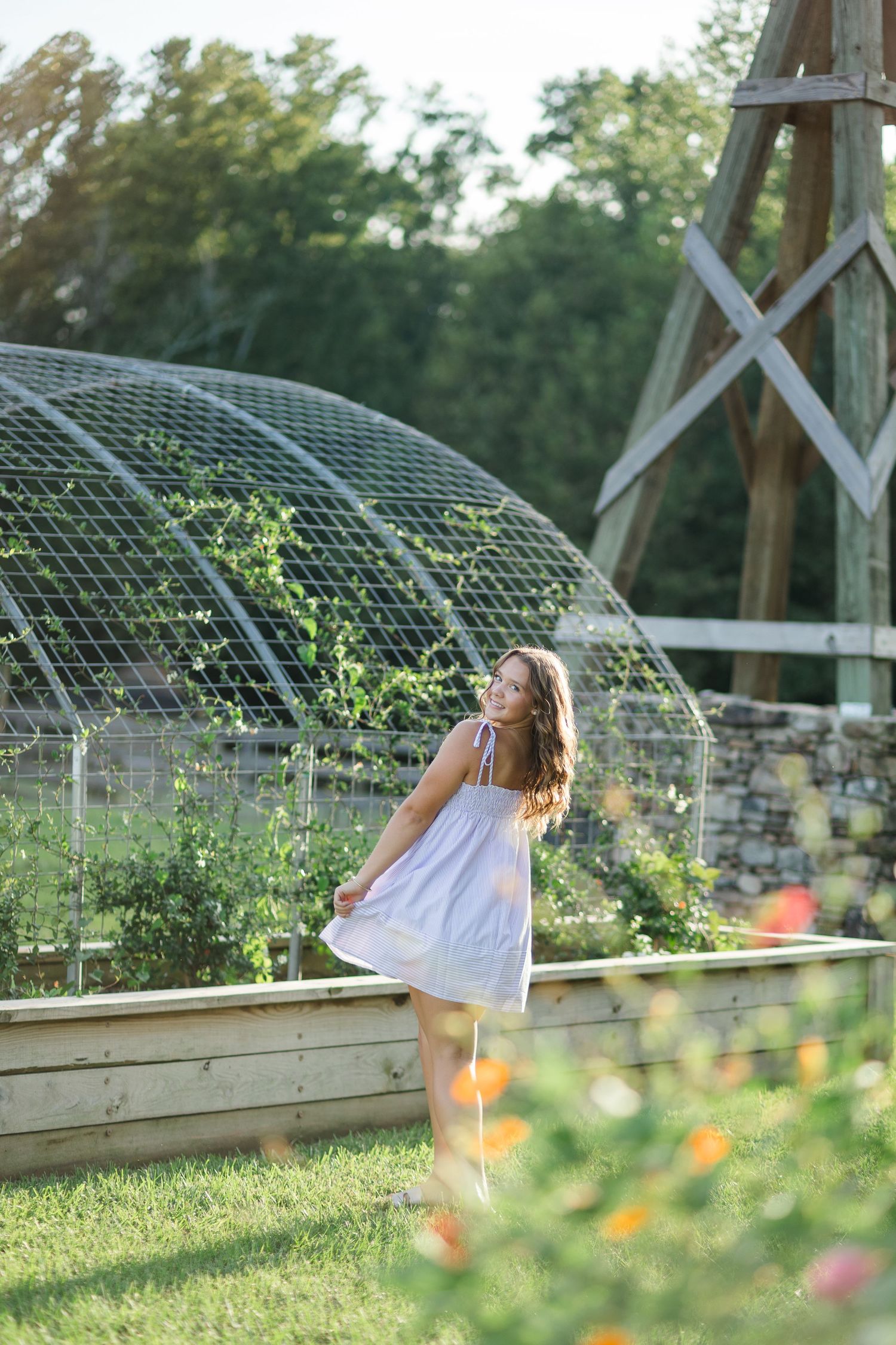 Young woman in white dress smiling in a lush garden with raised beds and a dome greenhouse structure.