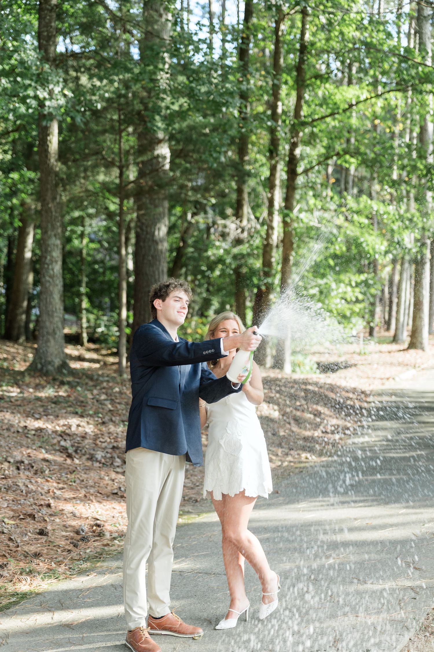 Couple celebrating engagement by popping champagne bottle outdoors in wooded setting, woman in white dress and man in navy blazer.