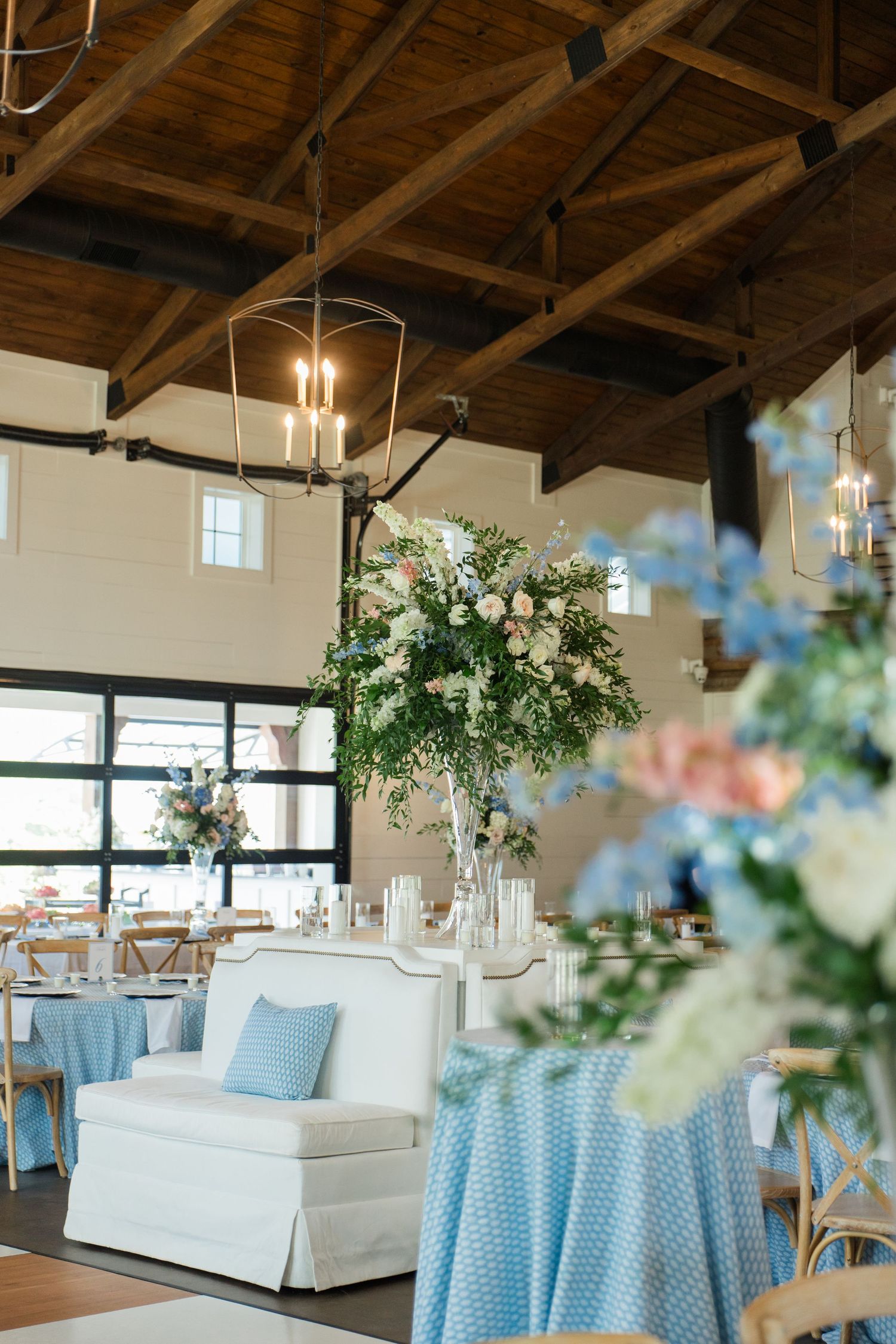 Elegant wedding reception venue with blue tablecloths, tall floral centerpieces, white sofas, and rustic wood beam ceiling.