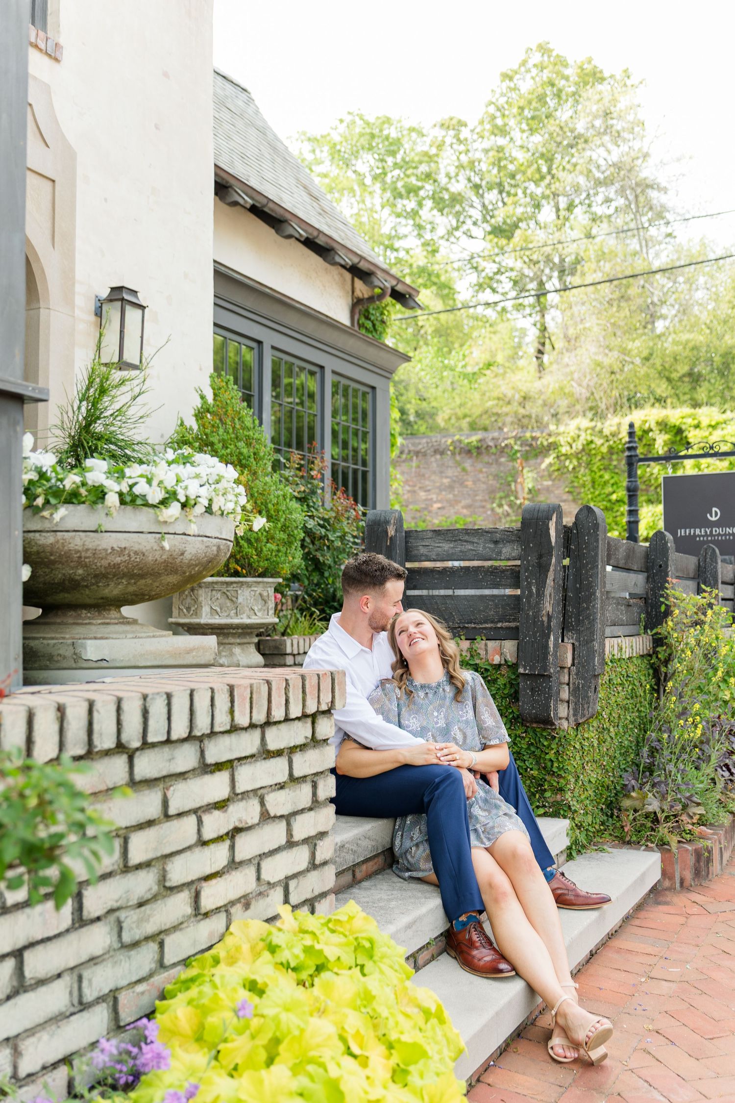 Happy couple sitting on stone steps surrounded by lush garden flowers during romantic engagement photos.
