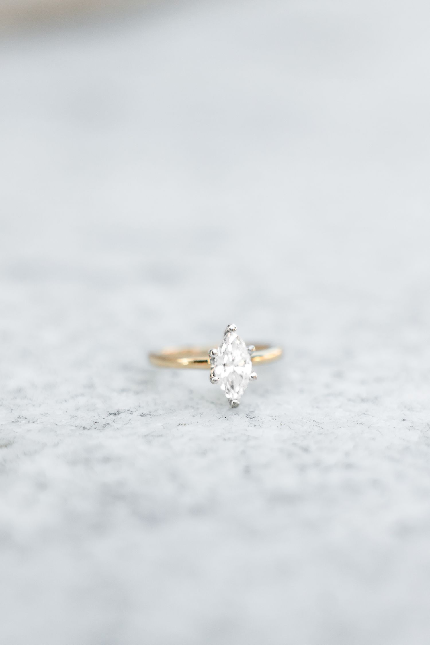 Gold solitaire engagement ring featuring a marquise-cut diamond on a light gray stone surface.