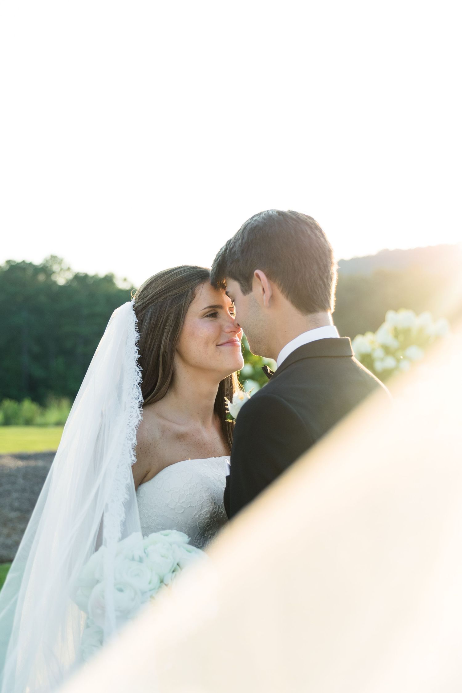 A bride and groom share a romantic moment outdoors at golden hour, with her white veil flowing in the sunlight.