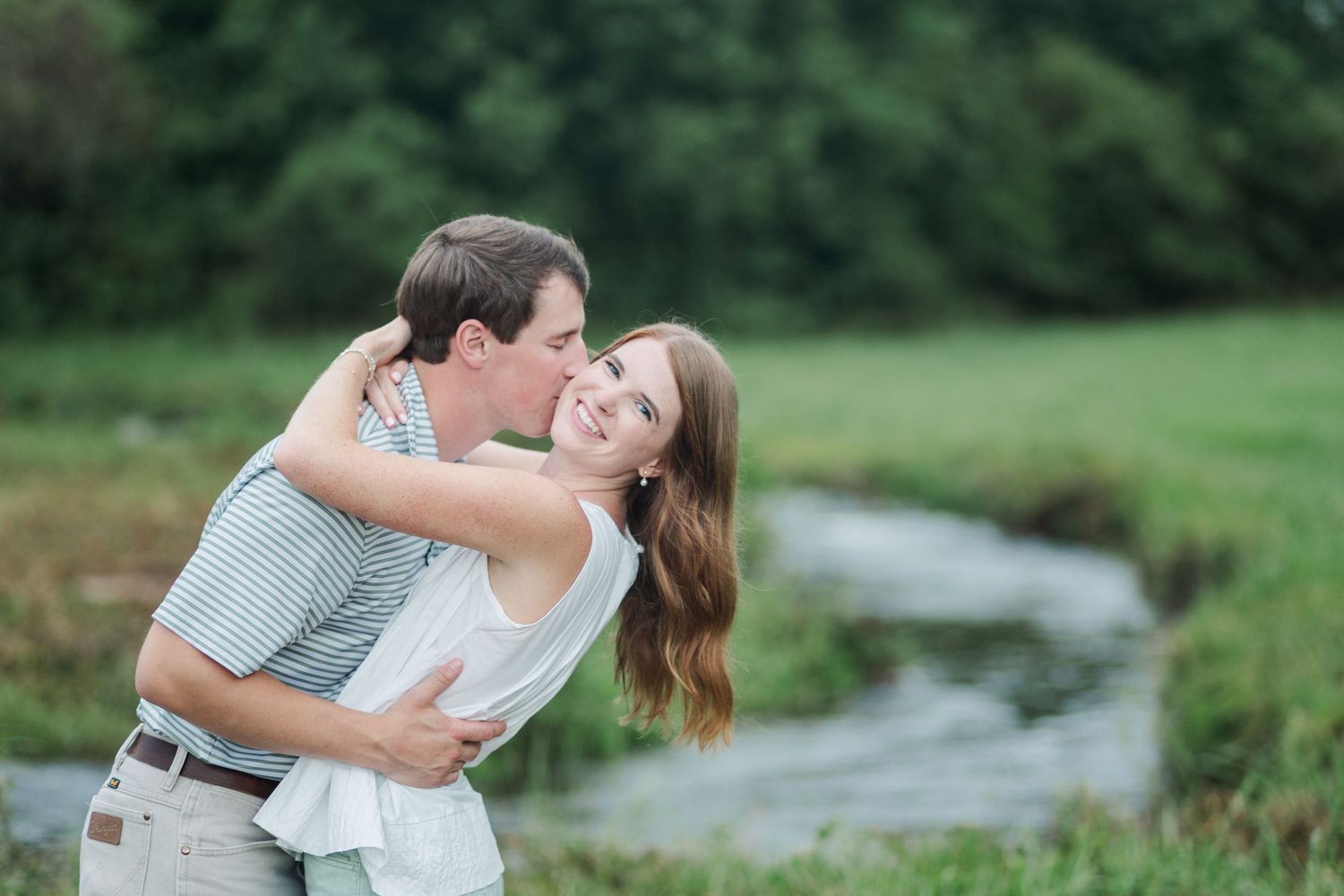 Man hugging and kissing smiling woman near a serene creek, both in casual summer outfits in a green meadow.