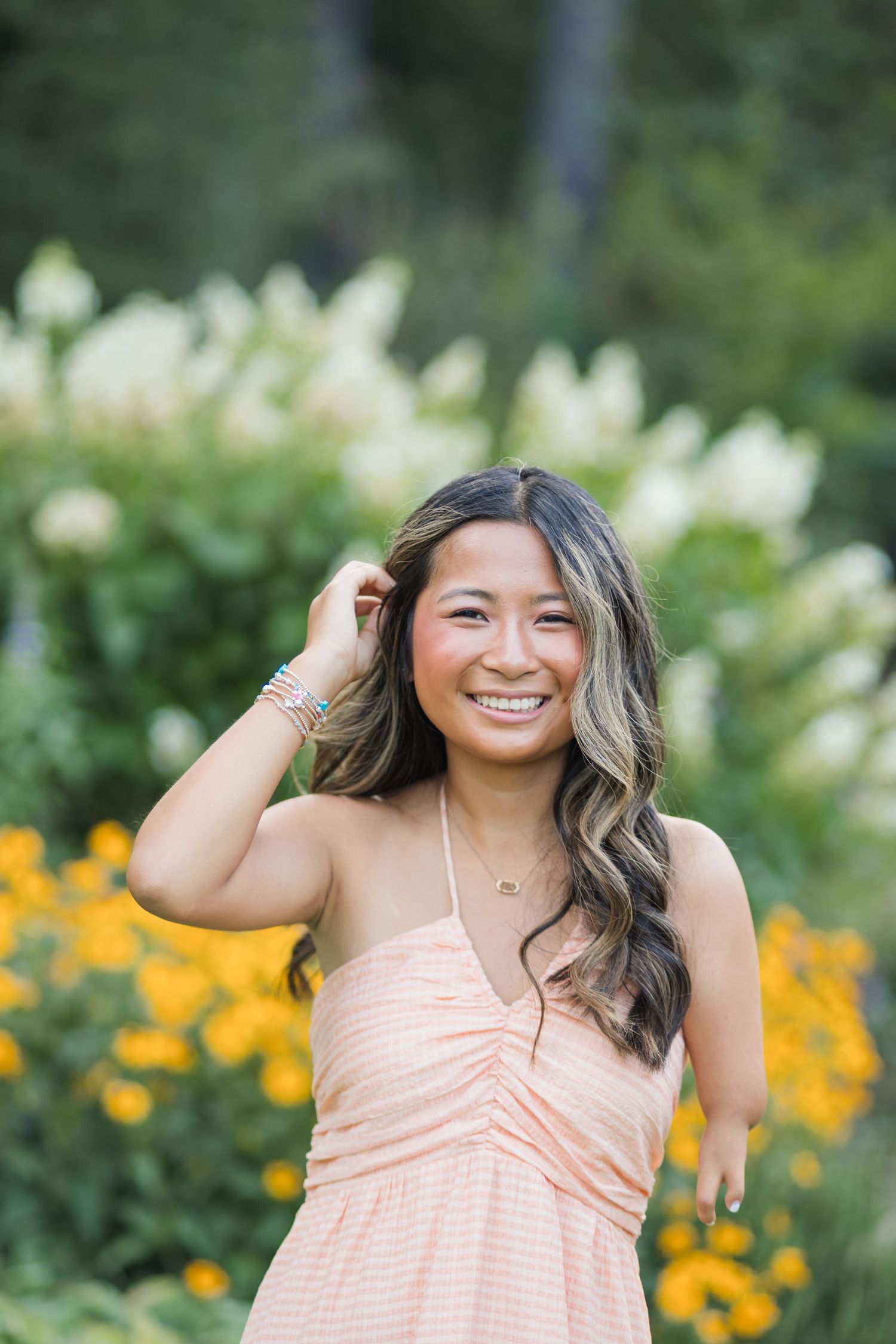 Smiling young woman in peach dress posing among yellow and white flowers in a garden.