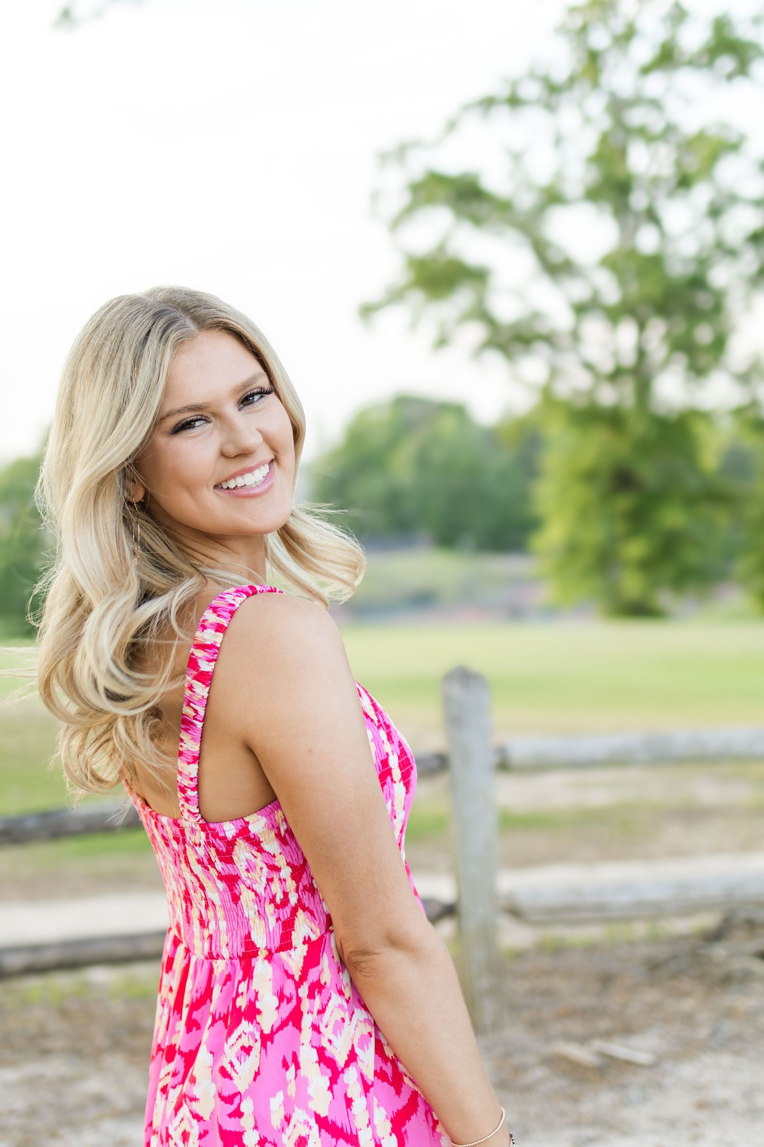 Smiling blonde woman in pink floral dress posing outdoors near a wooden fence.