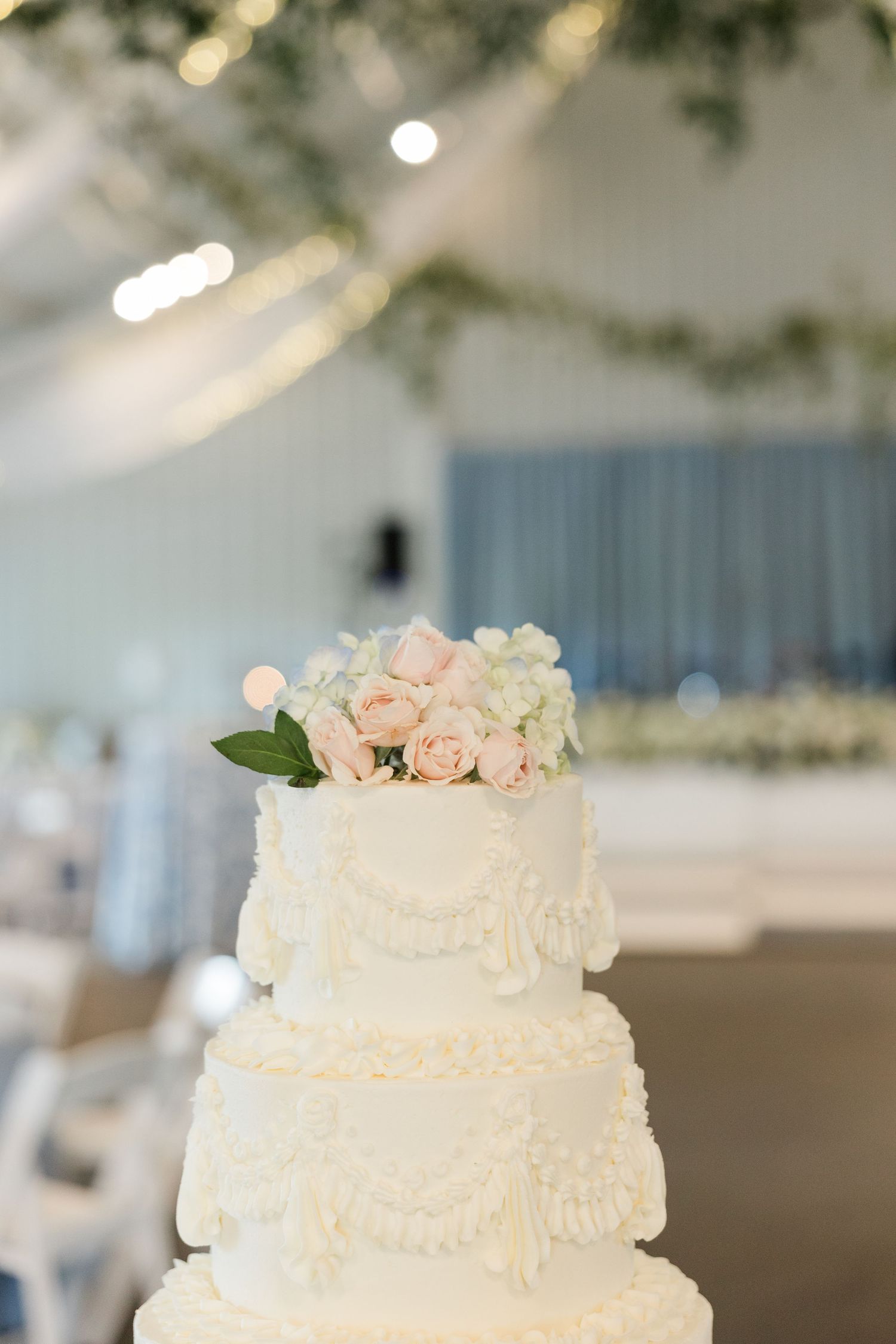 Elegant three-tier white wedding cake topped with blush pink roses and white hydrangeas in softly lit venue.