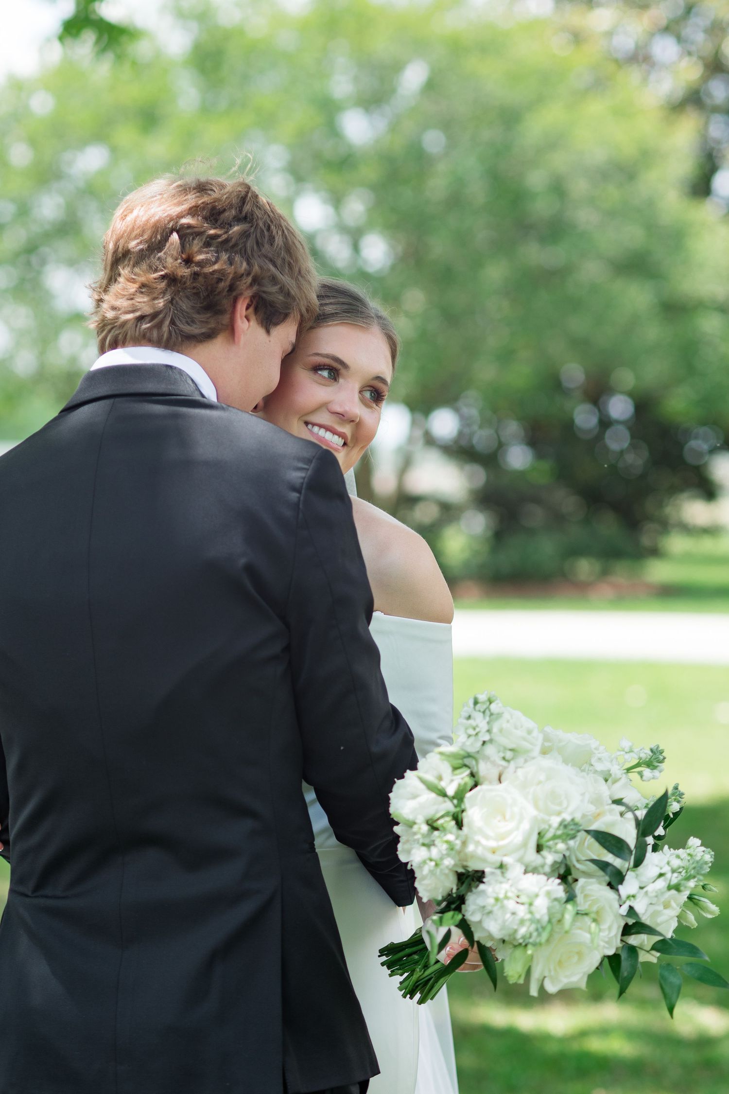 Bride and groom embrace outdoors, bride holding white rose bouquet and smiling over her shoulder.