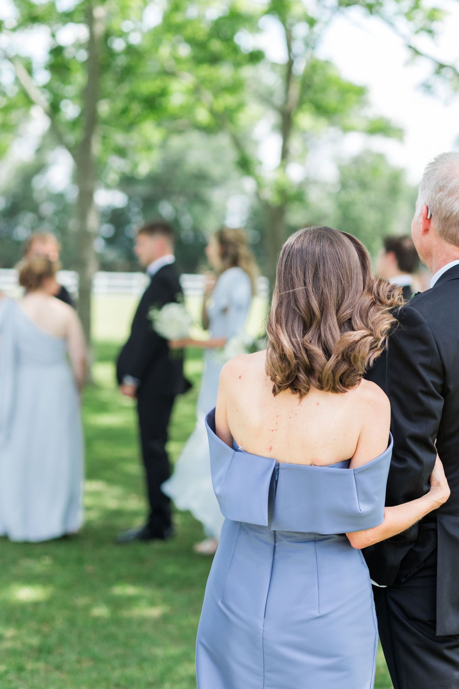 Mother of bride in blue off-shoulder dress watching outdoor wedding ceremony from behind.