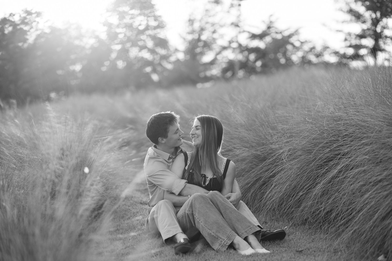 Black and white photo of a romantic couple sitting together in tall golden grass field at sunset.