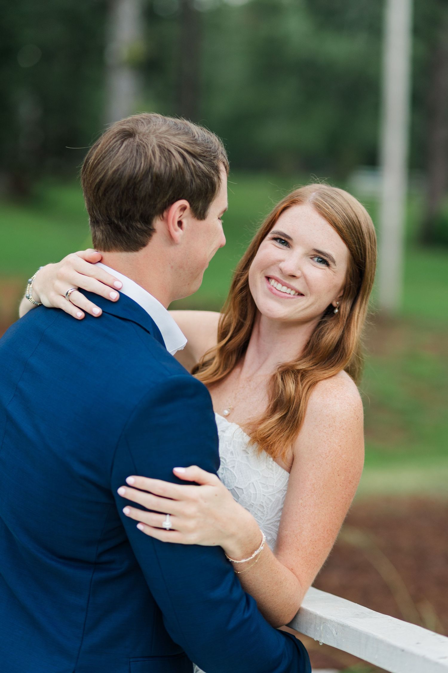 Smiling redhead bride embracing her groom in a blue suit against a white fence outdoors.