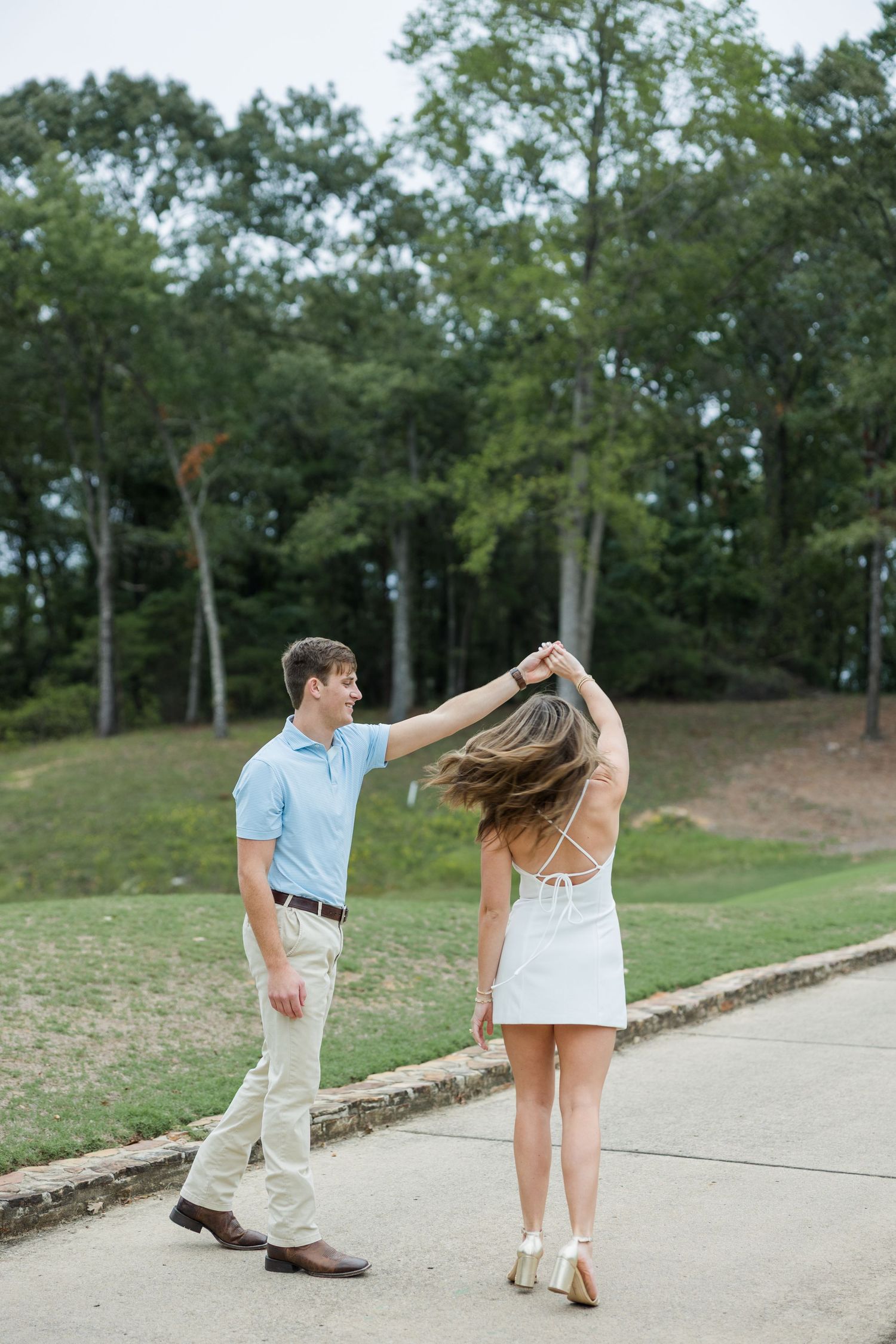 Man in a light blue polo twirls a woman in a white dress along a paved path beside a grassy hill and trees during an engagement shoot.
