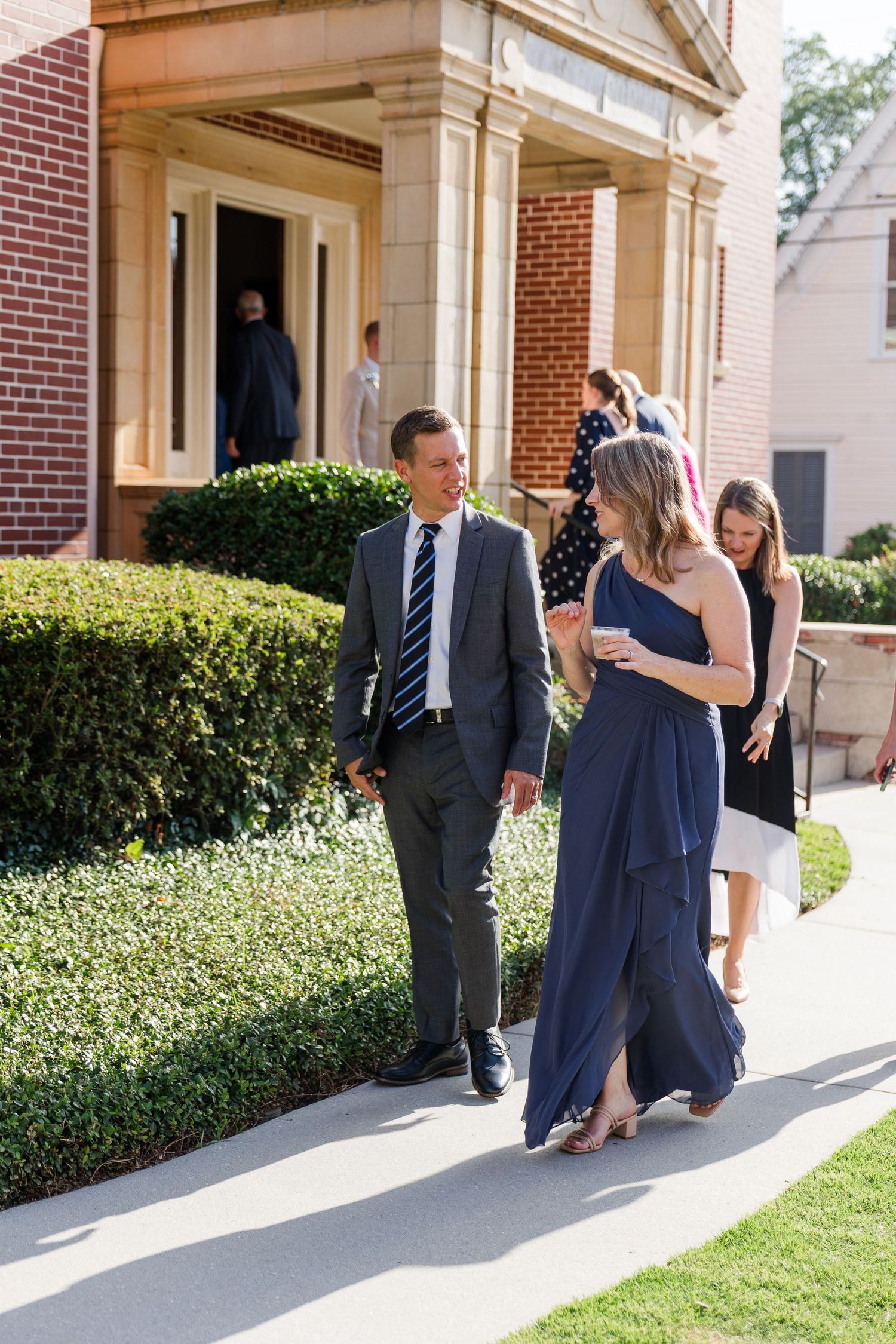 Wedding guests in formal attire walk along sidewalk outside brick church on sunny wedding day.