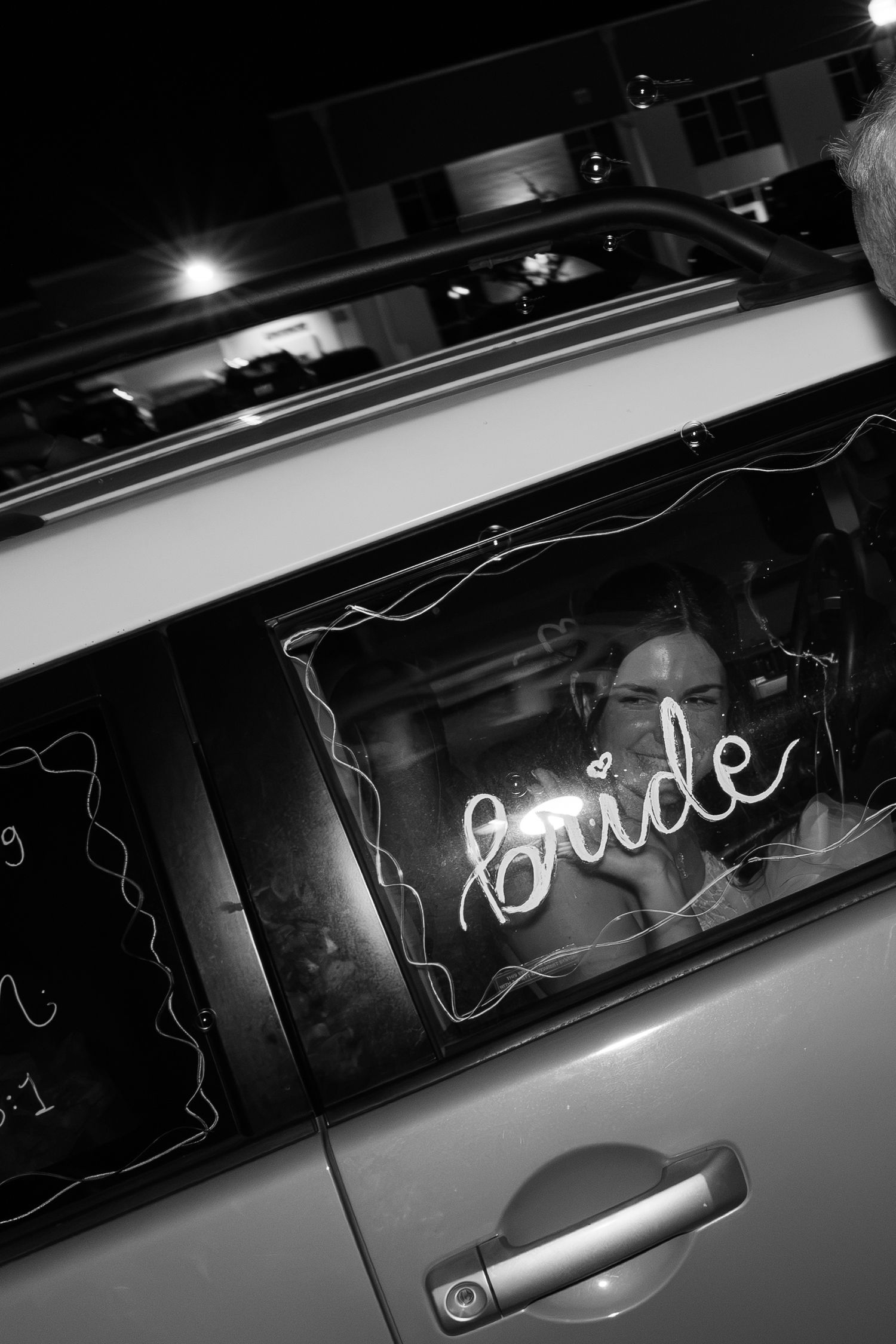 Black and white photo of bride sitting in car behind glowing neon bride sign at night.