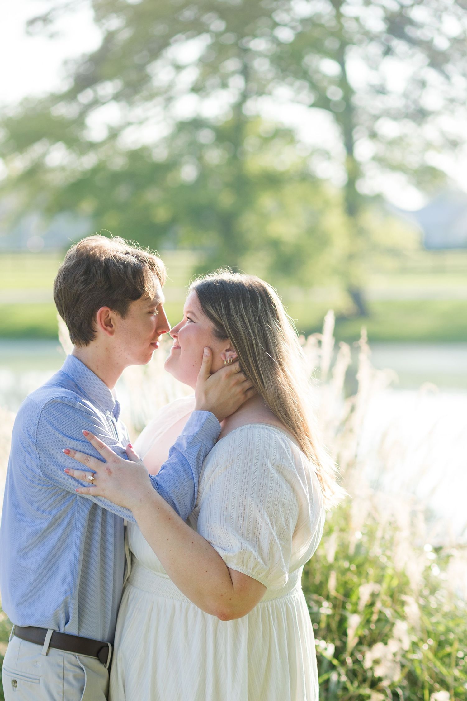 A couple shares a tender almost-kiss outdoors near a pond, surrounded by lush greenery in soft golden light.