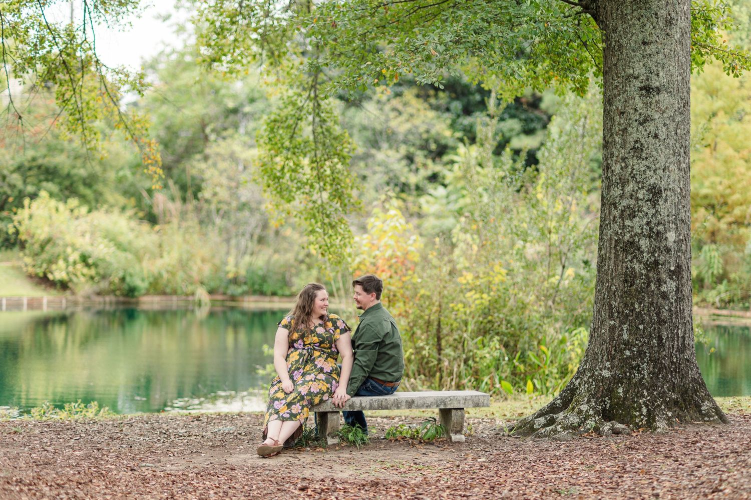 Couple sitting on a bench under a large tree by a serene pond, surrounded by lush greenery in an outdoor setting.
