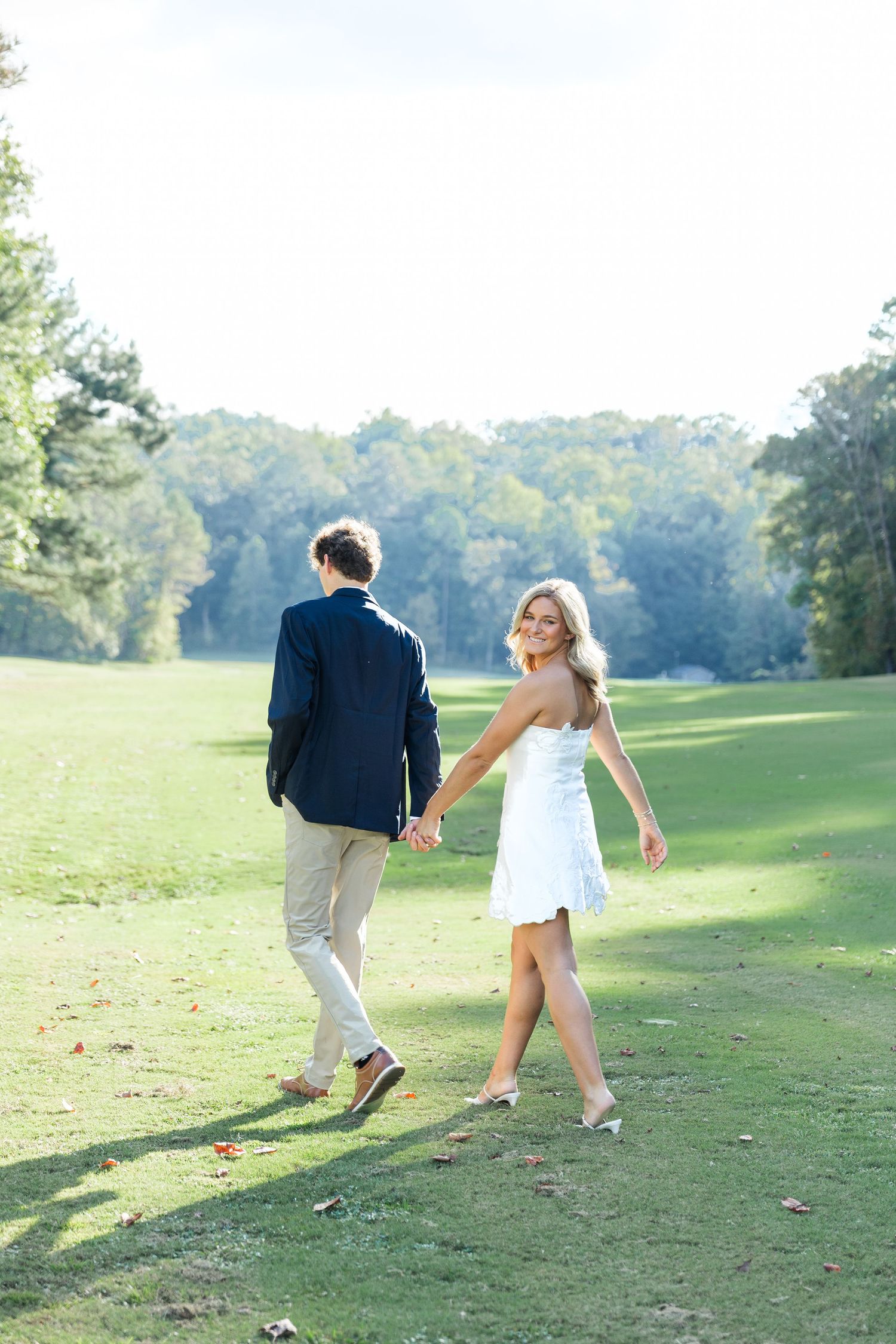 A smiling bride in a white dress holds hands with her groom in a navy suit while walking on a lush green lawn.